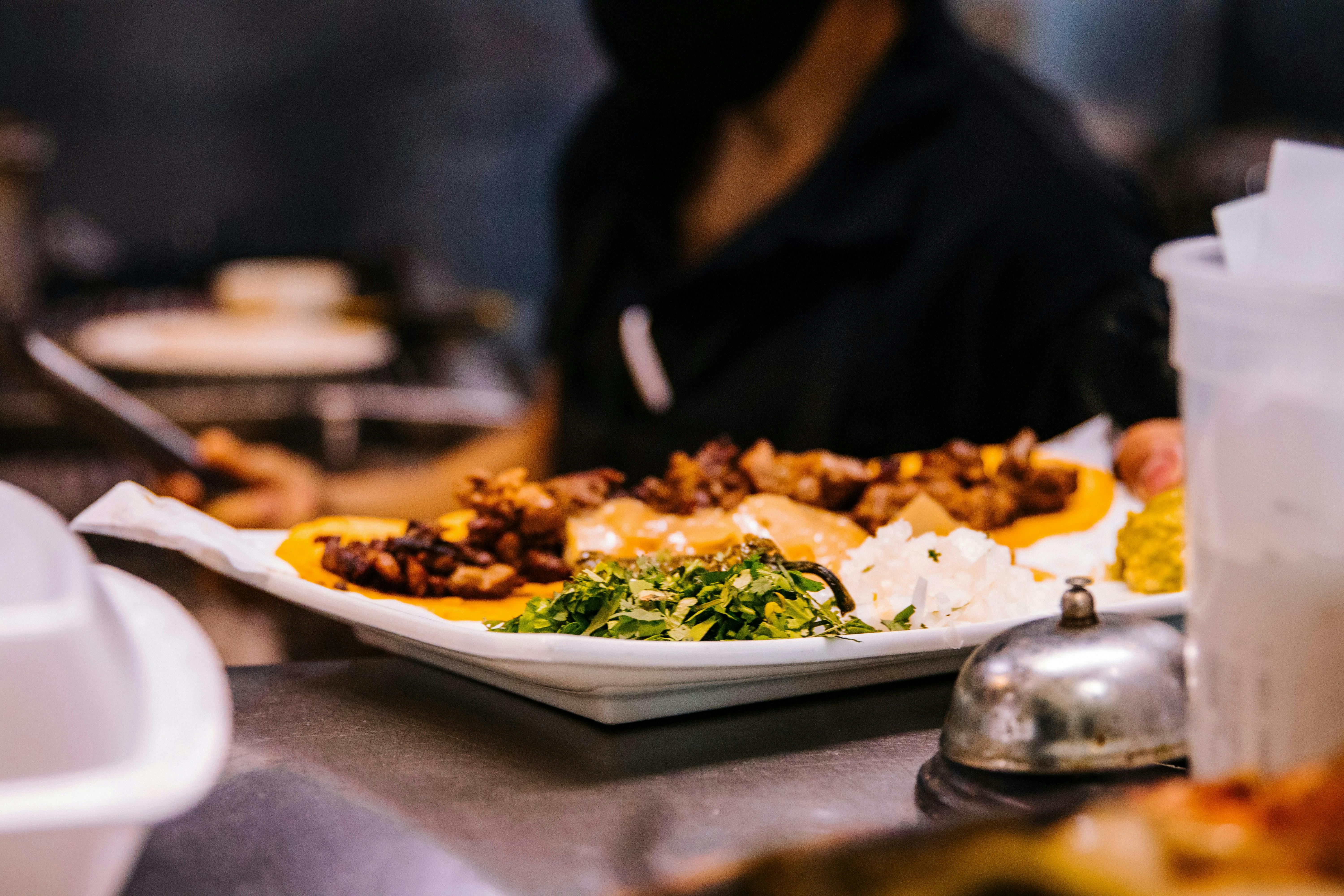 A close-up of a plate of street tacos on yellow corn tortillas, topped with meat, onions, and cilantro. A silver service bell sits in the immediate foreground on the counter, waiting for the order to be picked up.
