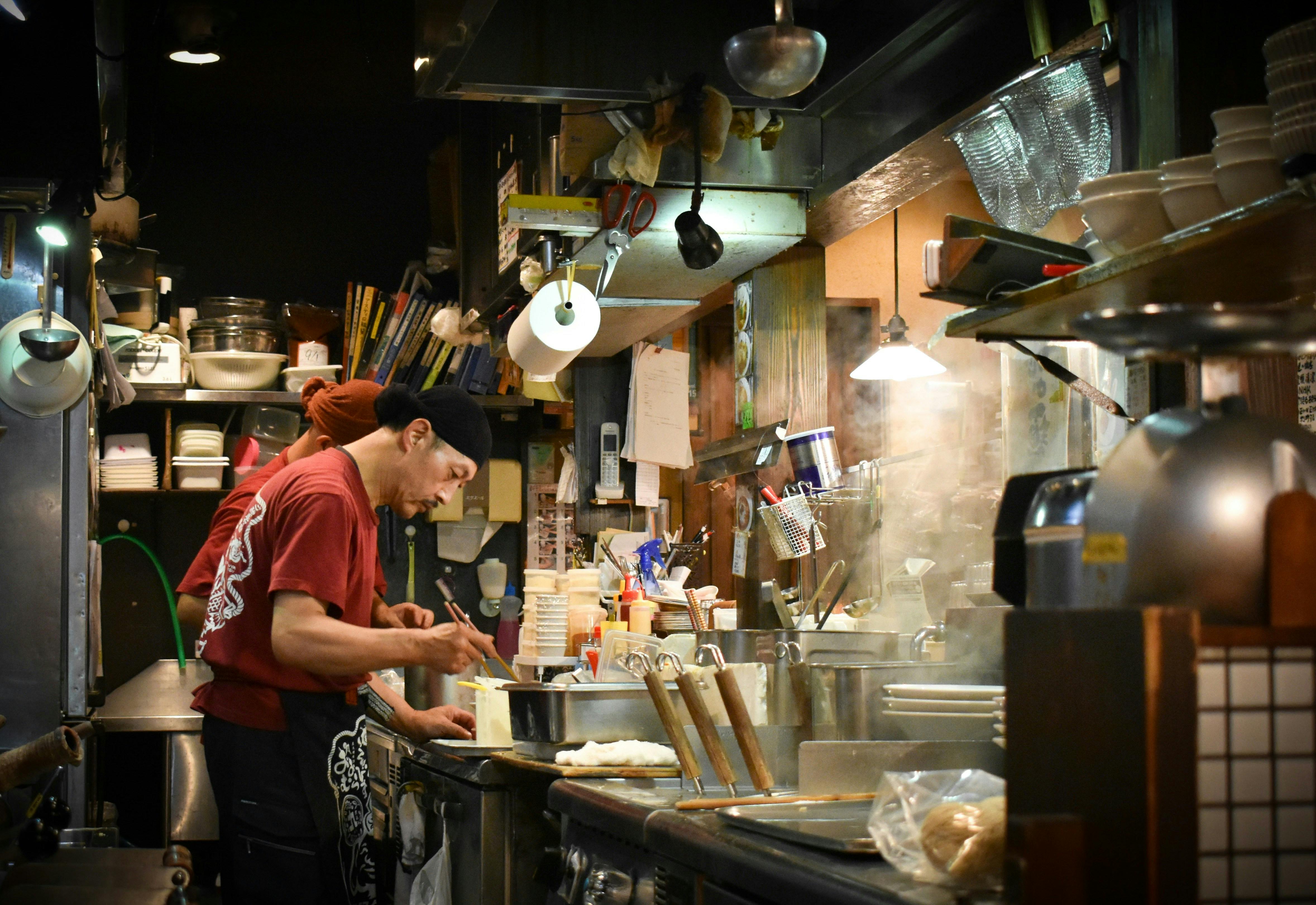 A chef in a red t-shirt and black head covering prepares food in a narrow, steamy commercial kitchen. The space is cluttered with shelves full of bowls, strainers, and cooking utensils, creating an authentic ramen shop atmosphere.

