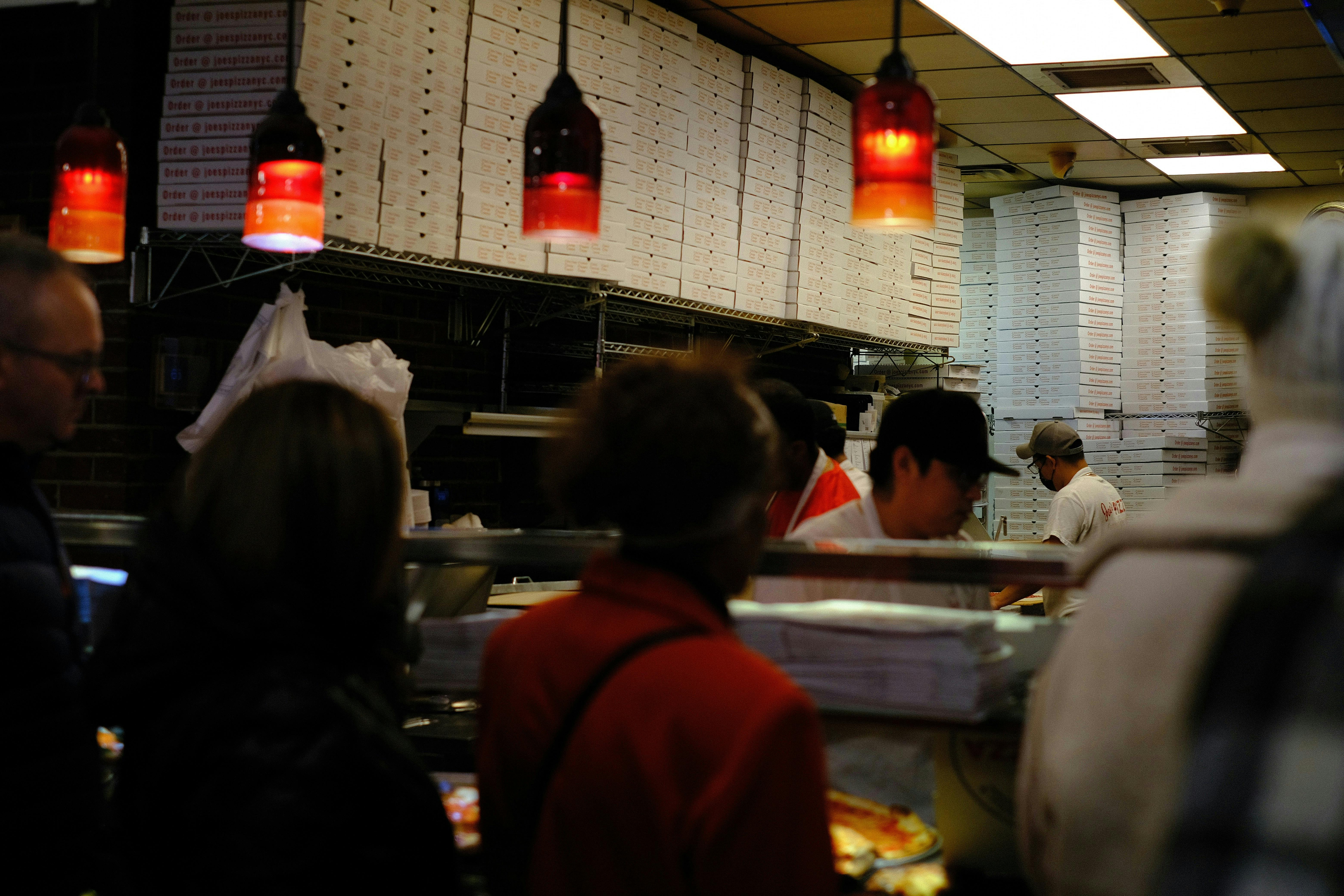 The interior of a busy pizzeria featuring floor-to-ceiling stacks of white pizza boxes lining the back wall. Red pendant lights hang from the ceiling above a stainless steel counter where staff in white uniforms serve customers.

