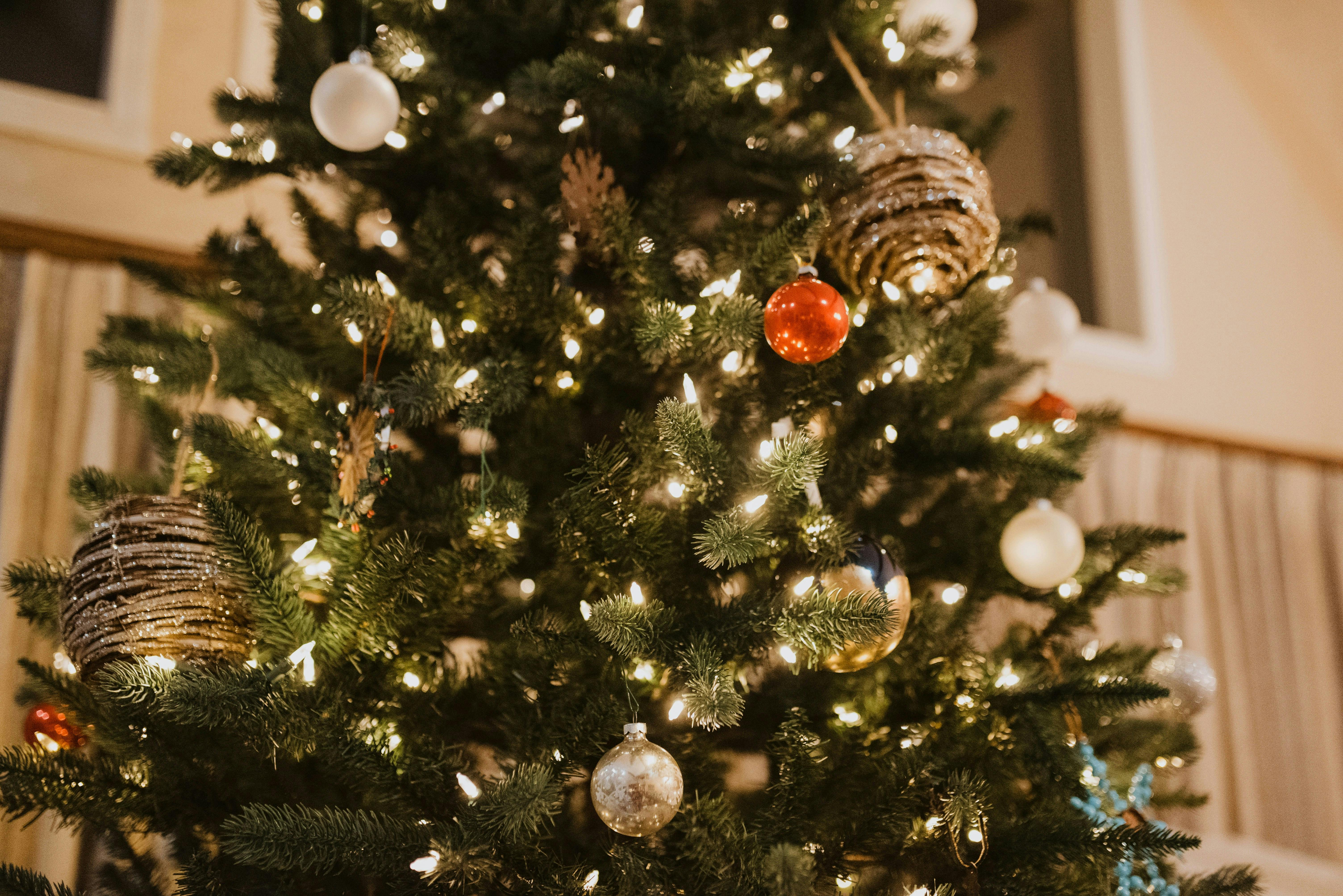 A brightly lit Christmas tree decorated with warm white fairy lights, red and silver baubles, and rustic twine-wrapped ornaments. The low-angle shot captures the cozy glow of the tree inside a home.