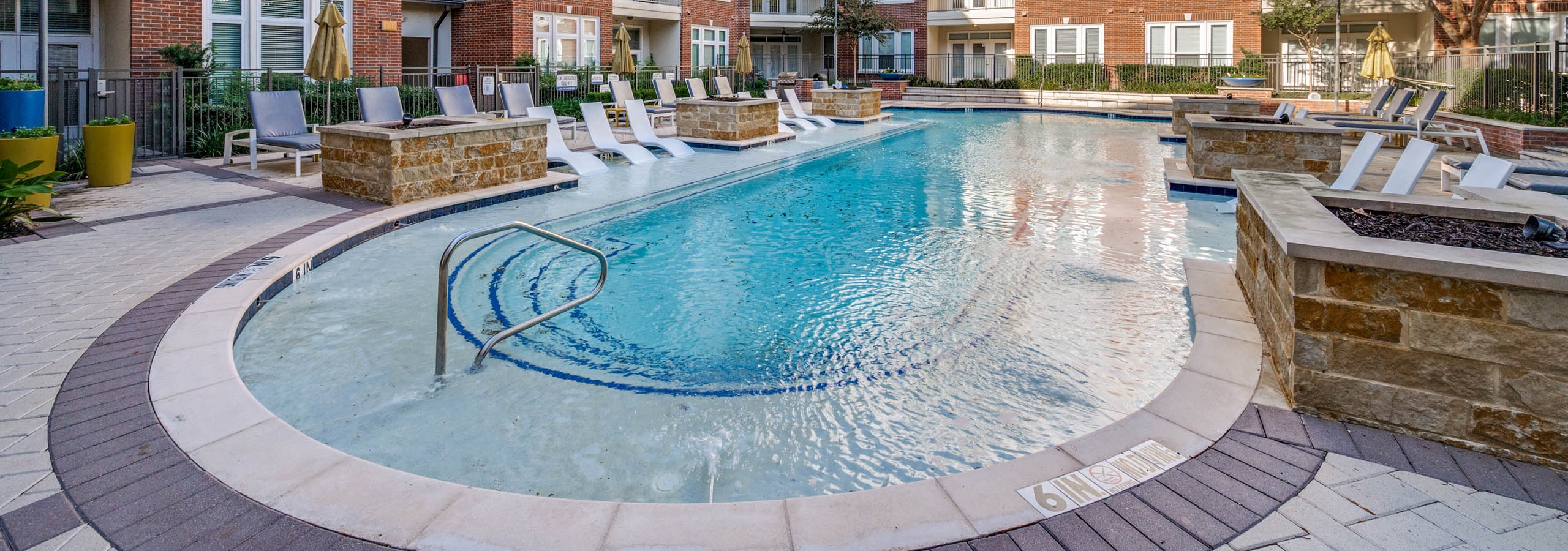 AMLI Frisco Crossing apartment daytime pool view with tanning ledge, lounge seating, and surrounding red brick buildings and trees.