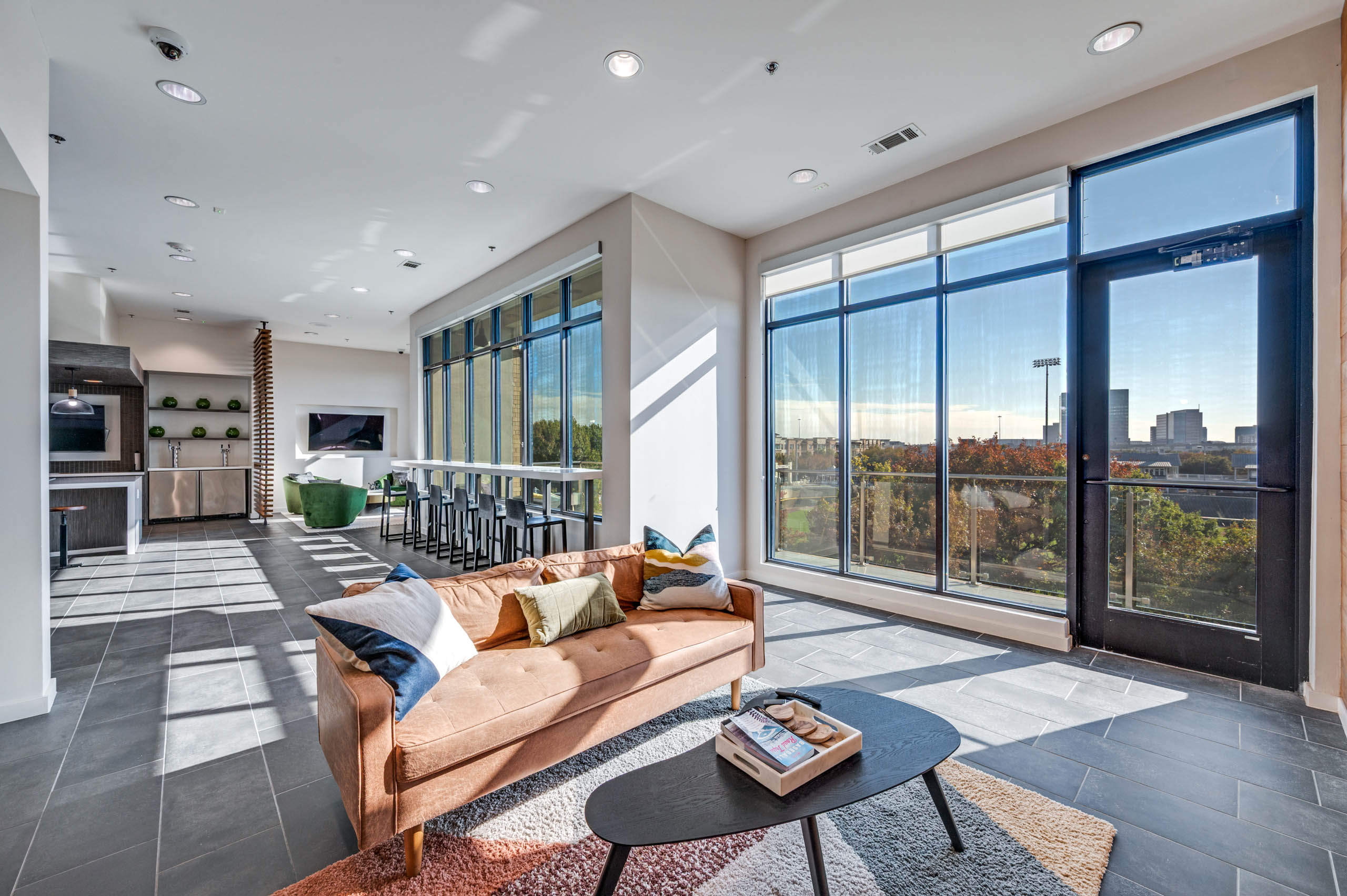 Spacious lounge area with large windows, bar seating, plush tan sofa over a decorative rug at AMLI Frisco Crossing apartment sky lounge.