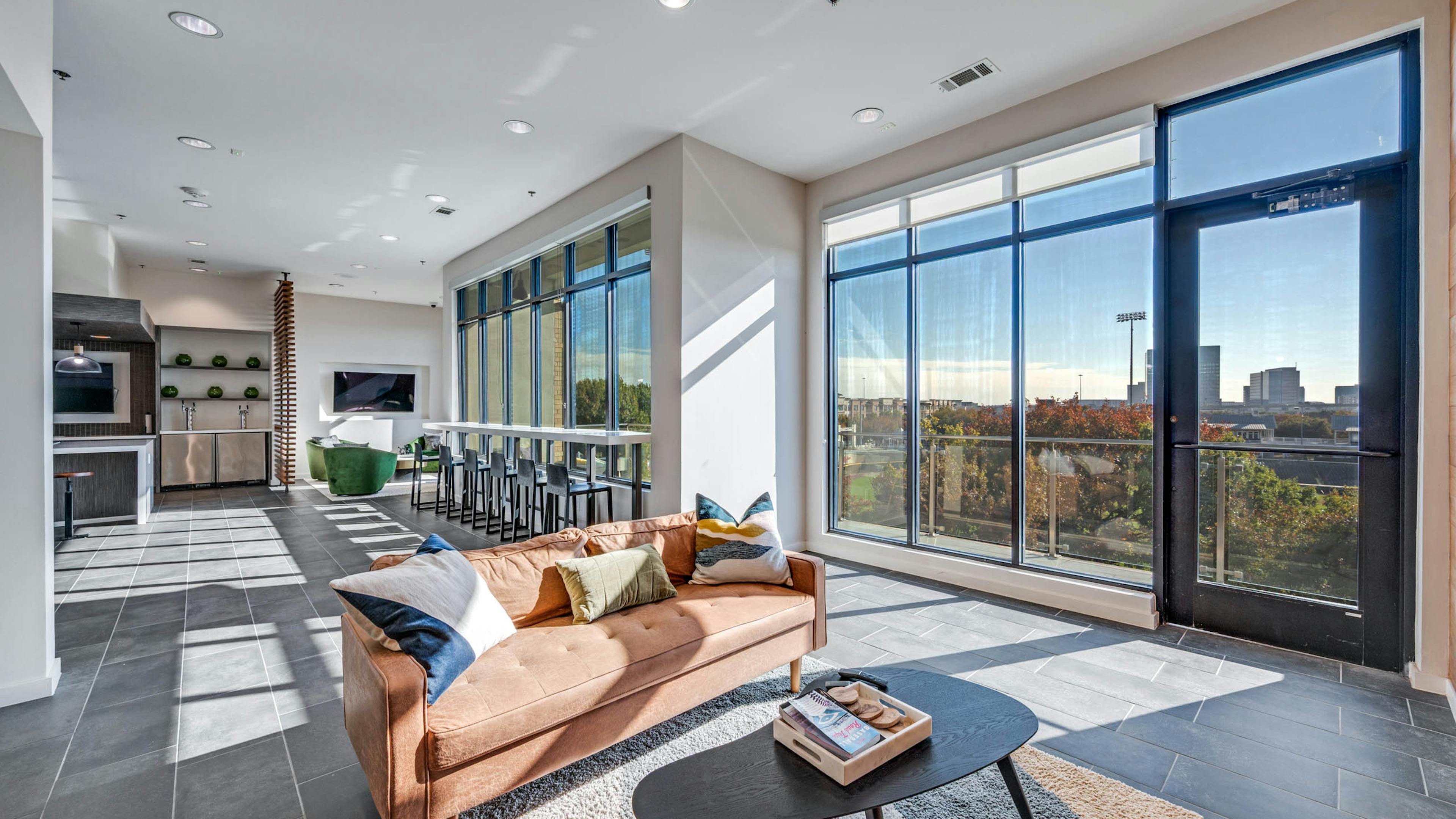 Spacious lounge area with large windows, bar seating, plush tan sofa over a decorative rug at AMLI Frisco Crossing apartment sky lounge.