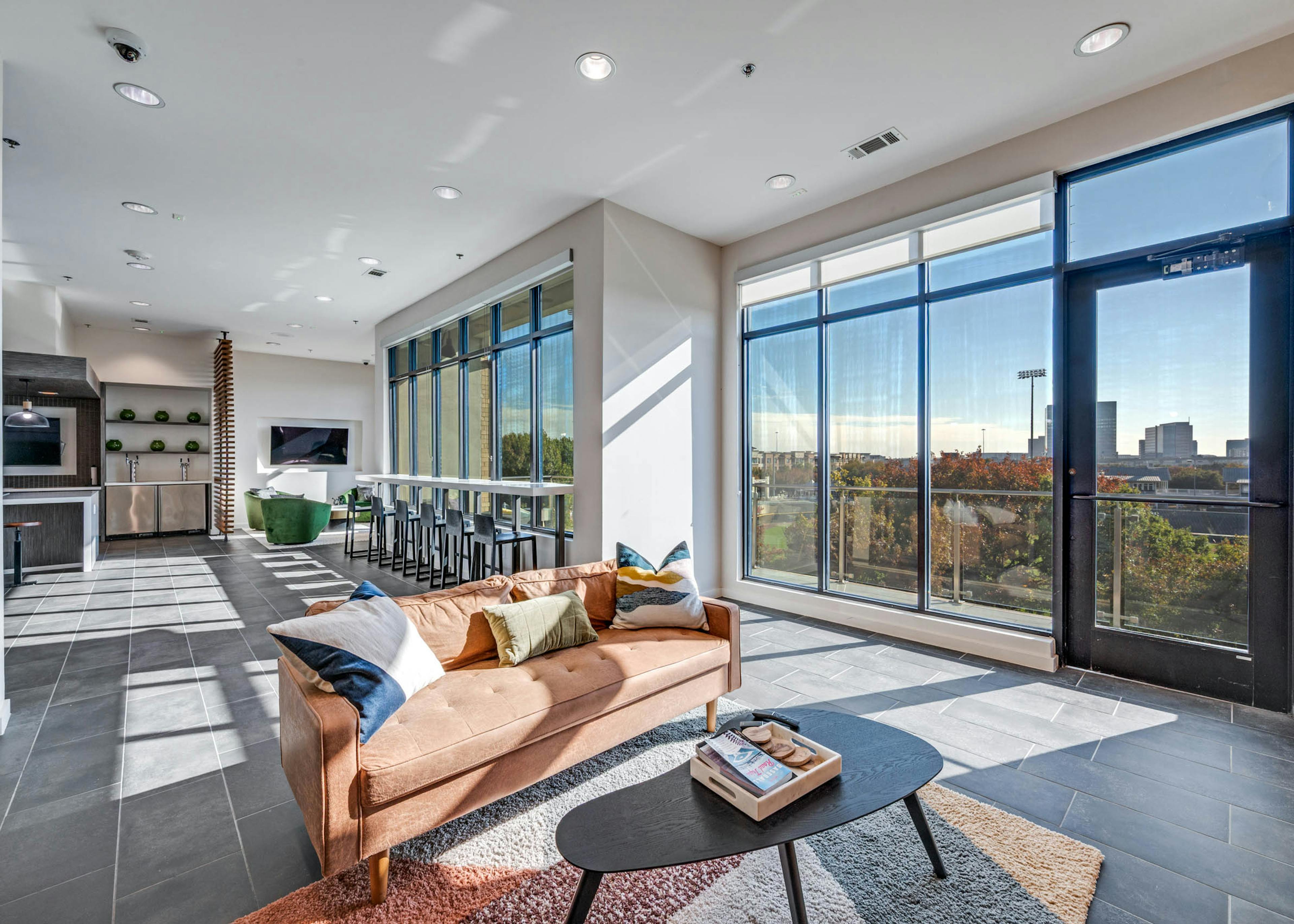 Spacious lounge area with large windows, bar seating, plush tan sofa over a decorative rug at AMLI Frisco Crossing apartment sky lounge.