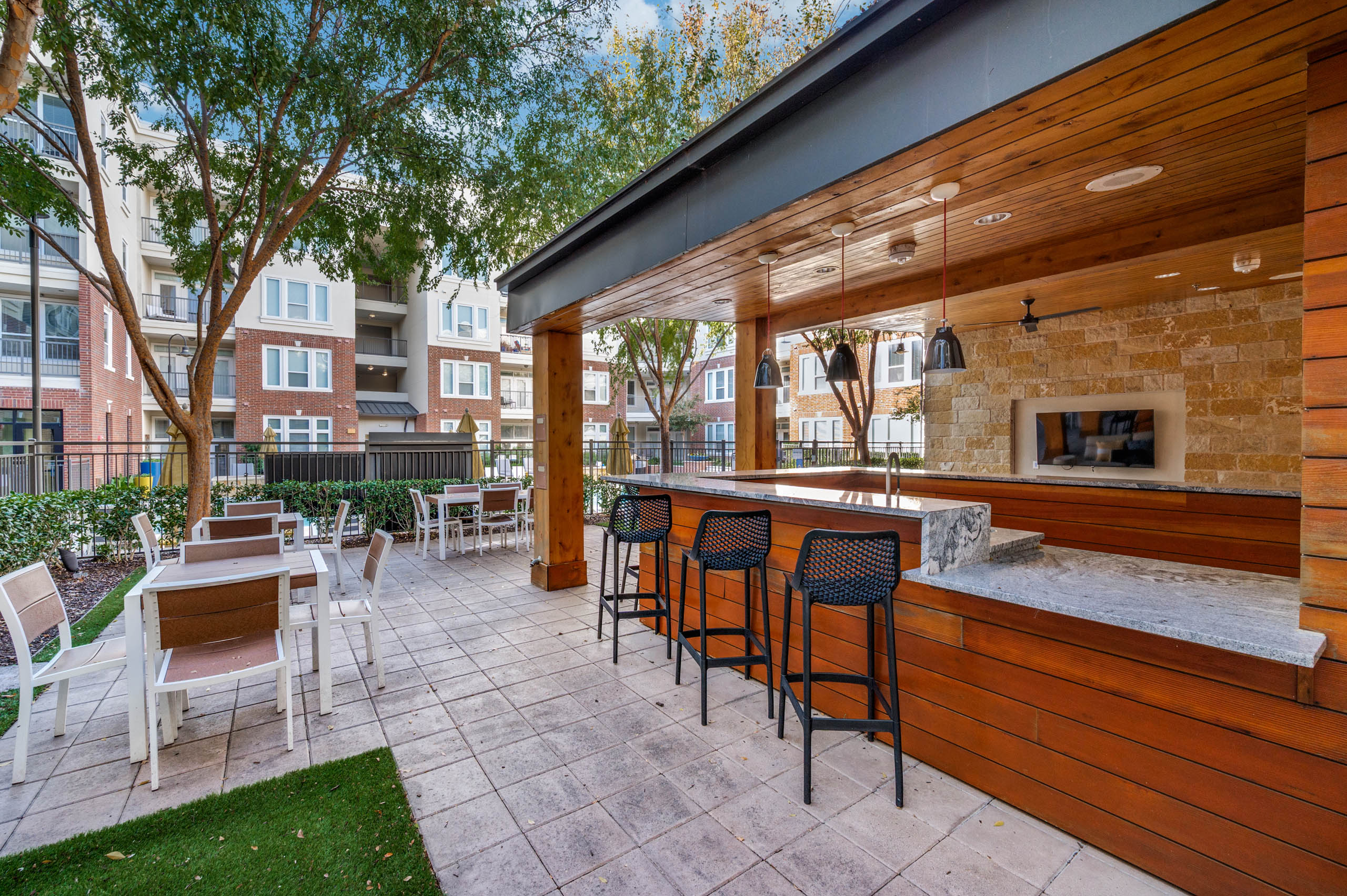 Covered outdoor lounge with bar seating, pendant lights and flat screen TV at AMLI Frisco Crossing apartments, with tables and trees nearby