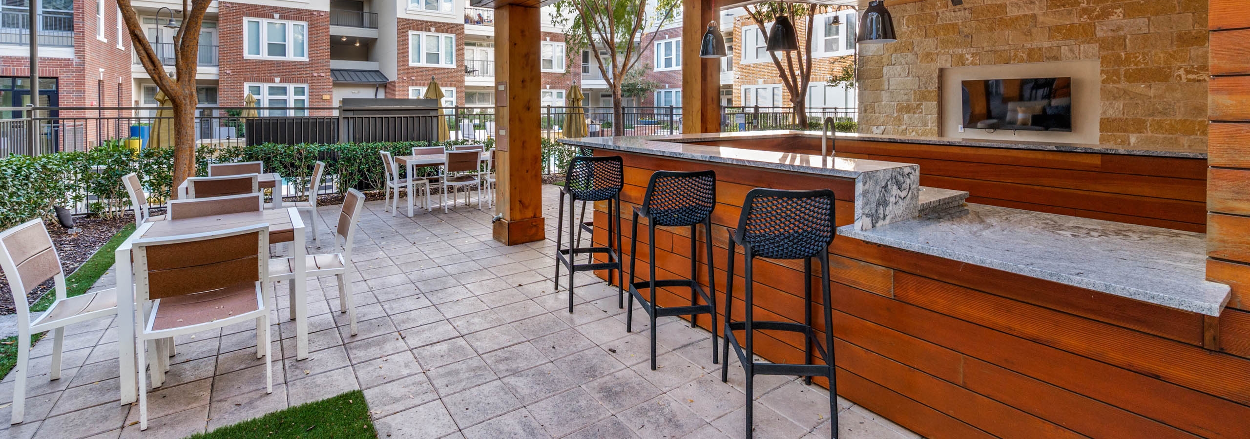 Covered outdoor lounge with bar seating, pendant lights and flat screen TV at AMLI Frisco Crossing apartments, with tables and trees nearby