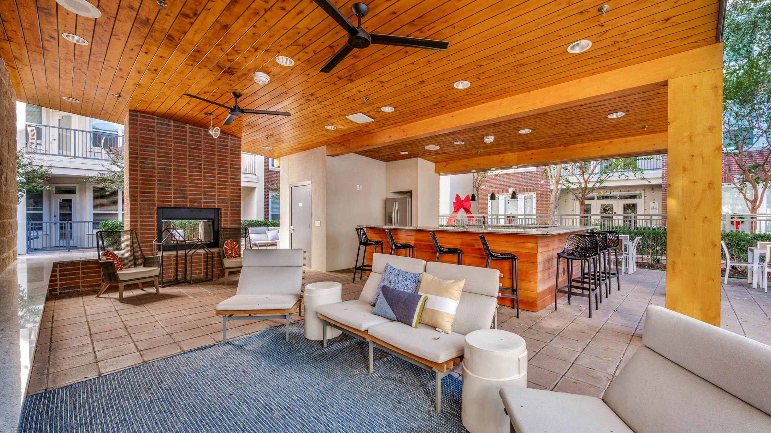 Shaded outdoor lounge area with ceiling fans, cushioned seating, brick fireplace, and bar at AMLI Frisco Crossing apartments