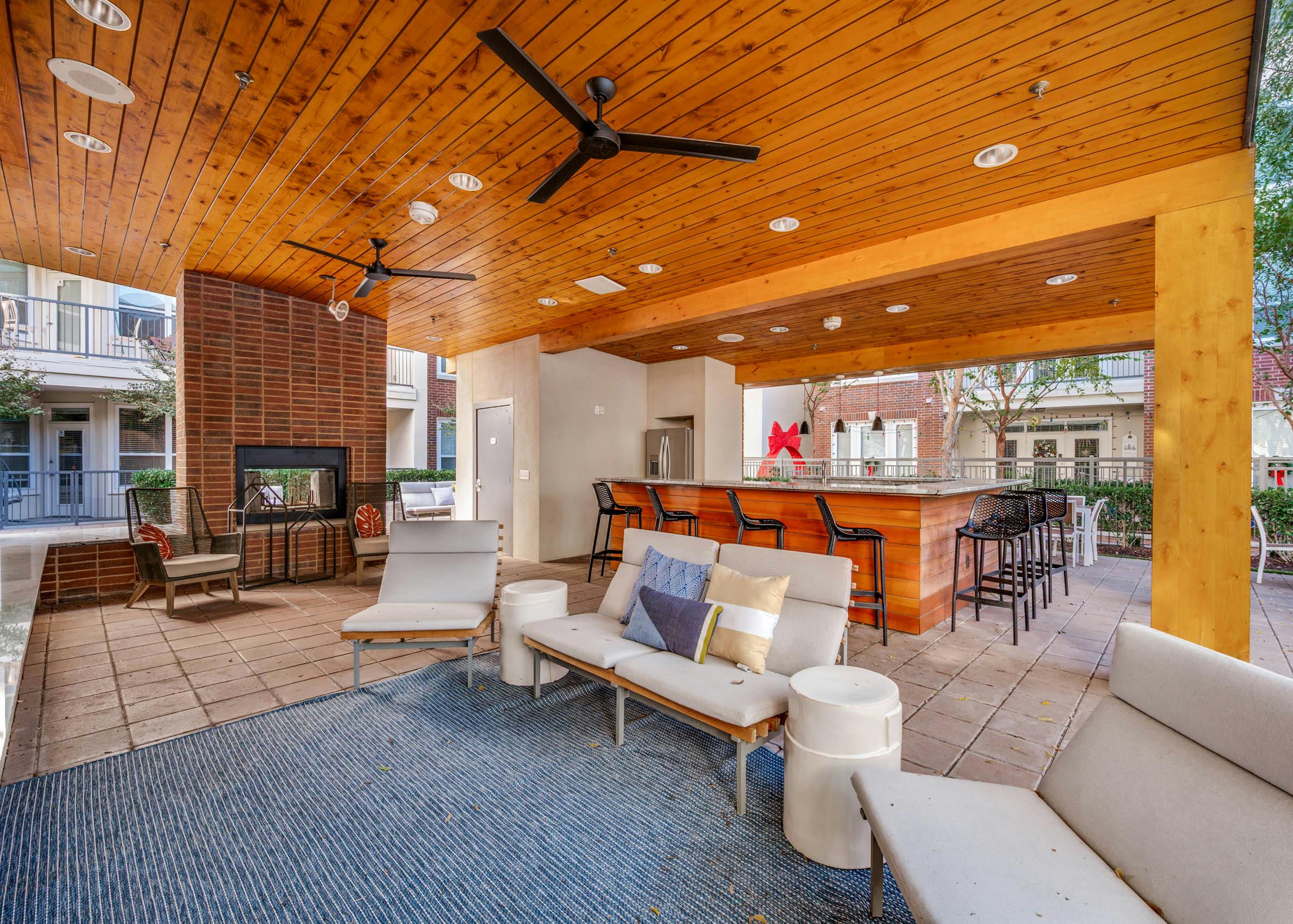 Shaded outdoor lounge area with ceiling fans, cushioned seating, brick fireplace, and bar at AMLI Frisco Crossing apartments
