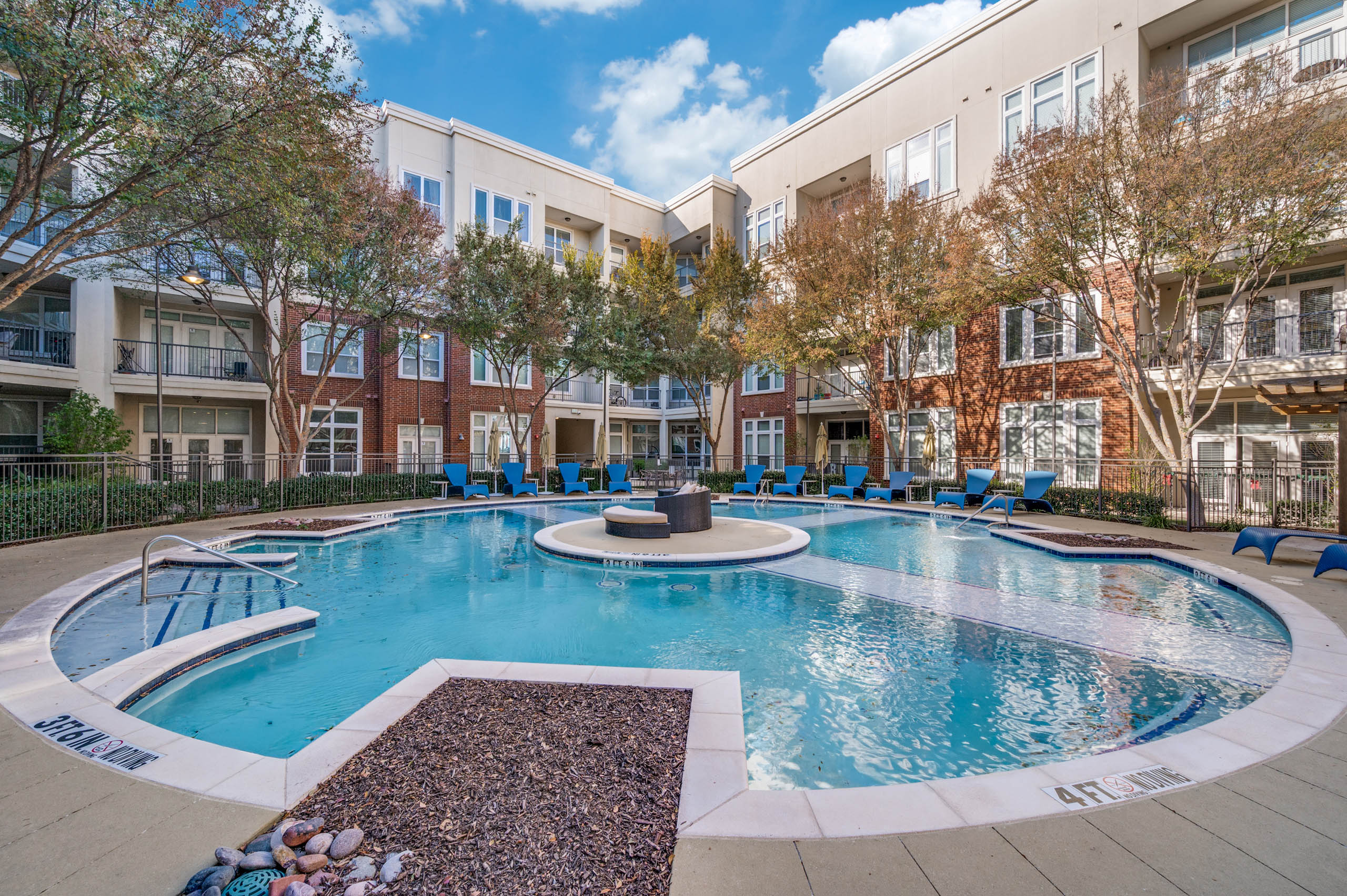Courtyard view of round resort-style pool with blue lounge chairs, trees, and red brick buildings at AMLI Frisco Crossing apartments.