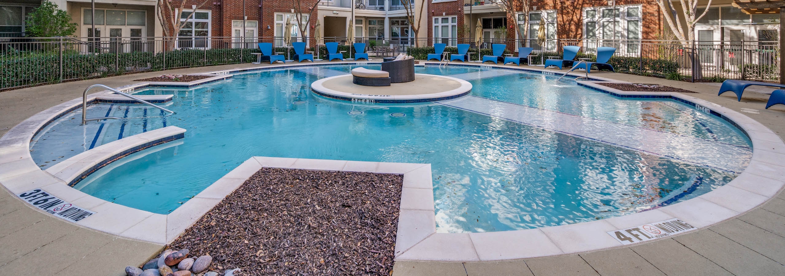 Courtyard view of round resort-style pool with blue lounge chairs, trees, and red brick buildings at AMLI Frisco Crossing apartments.