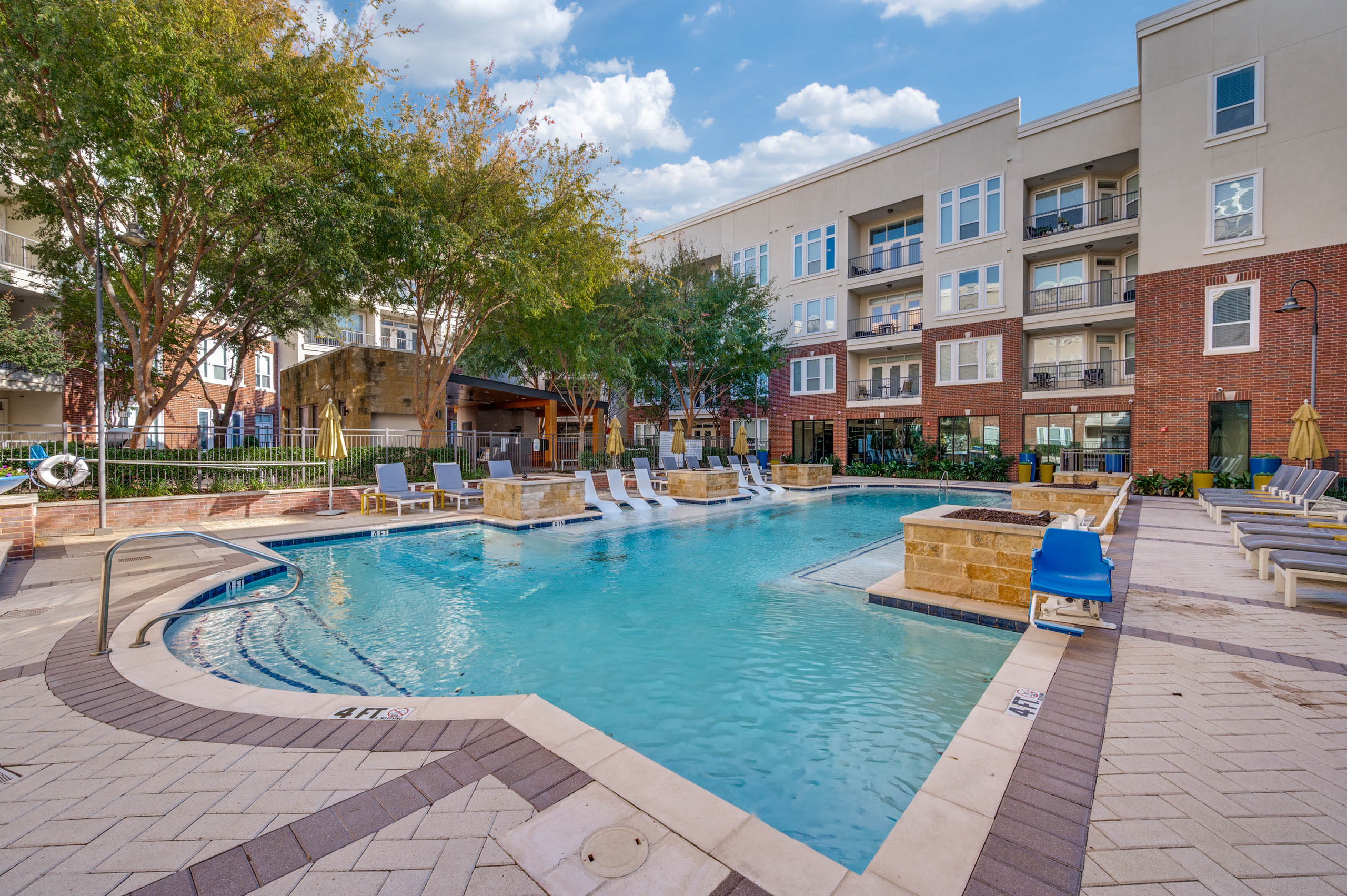 Resort-style swimming pool with white in-water loungers, umbrellas, cushioned seating and brick buildings at AMLI Frisco Crossing apartments.