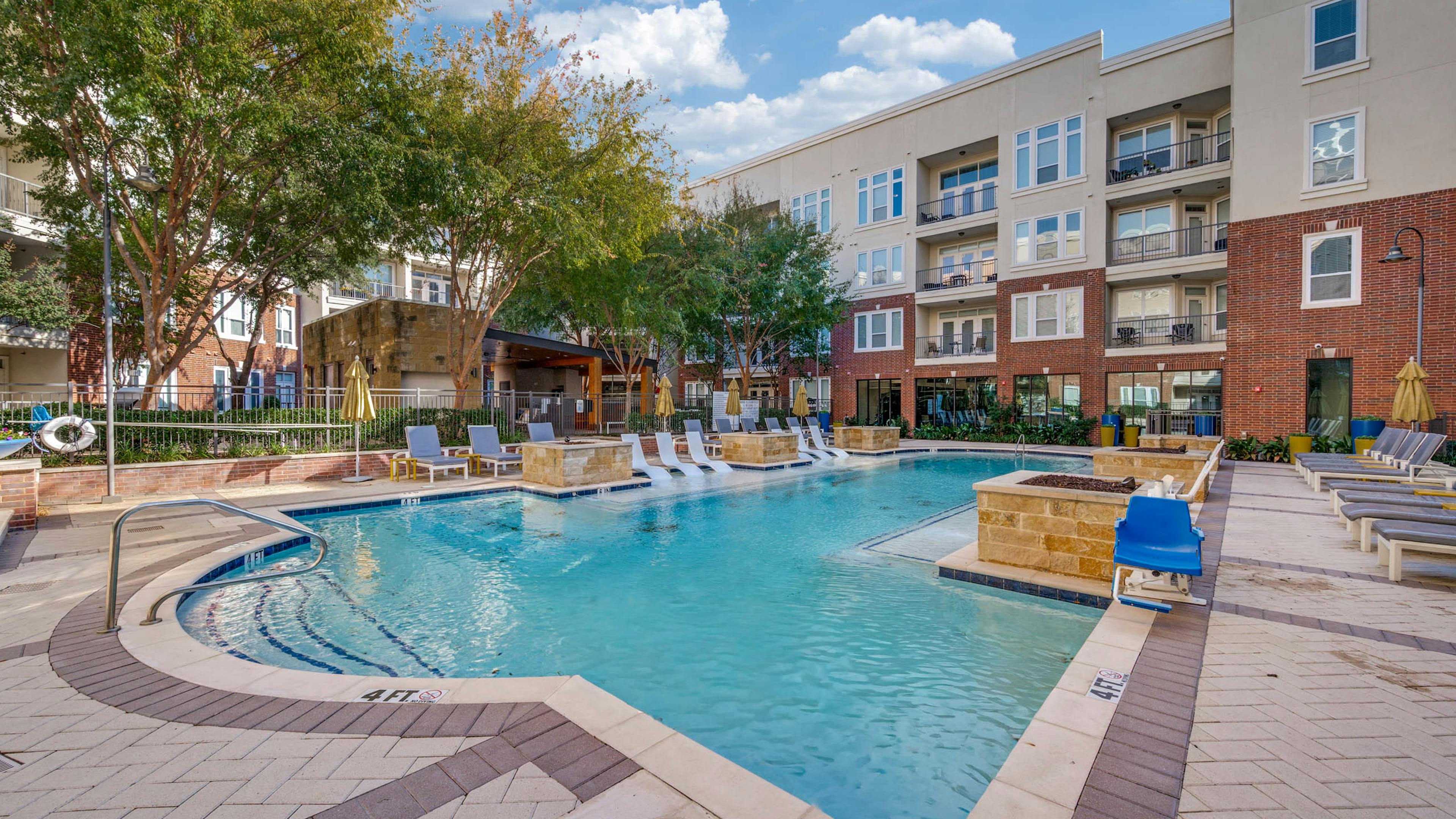 Resort-style swimming pool with white in-water loungers, umbrellas, cushioned seating and brick buildings at AMLI Frisco Crossing apartments.
