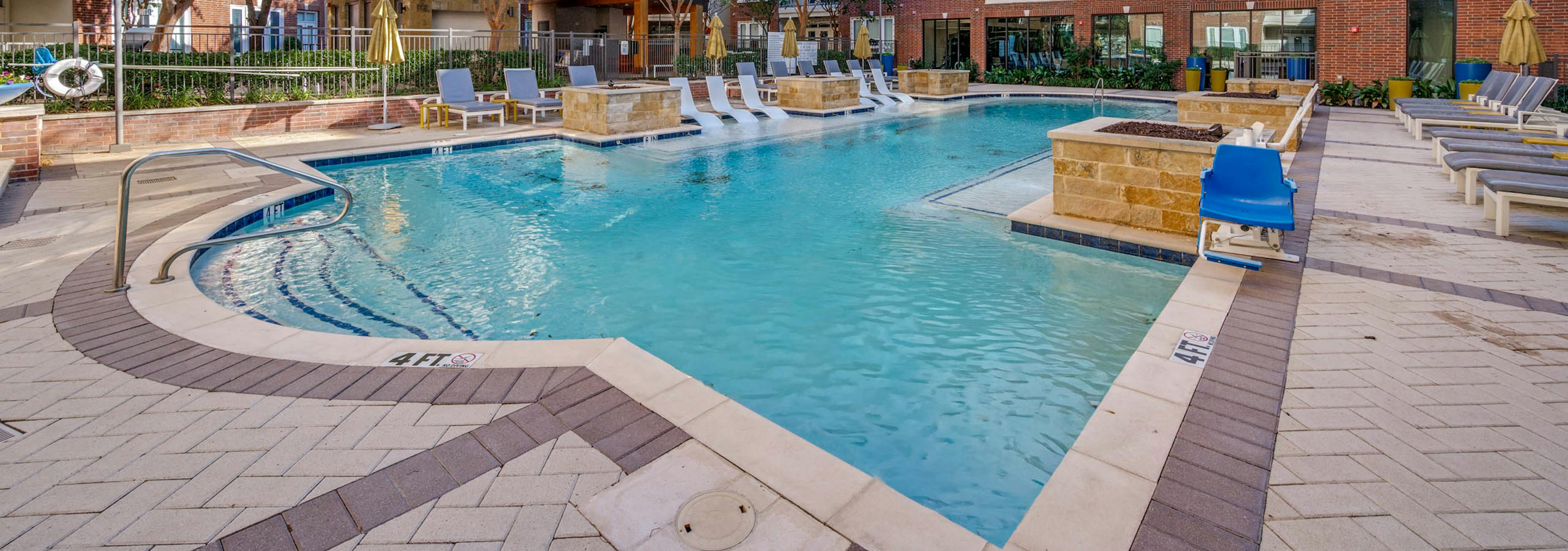 Resort-style swimming pool with white in-water loungers, umbrellas, cushioned seating and brick buildings at AMLI Frisco Crossing apartments.