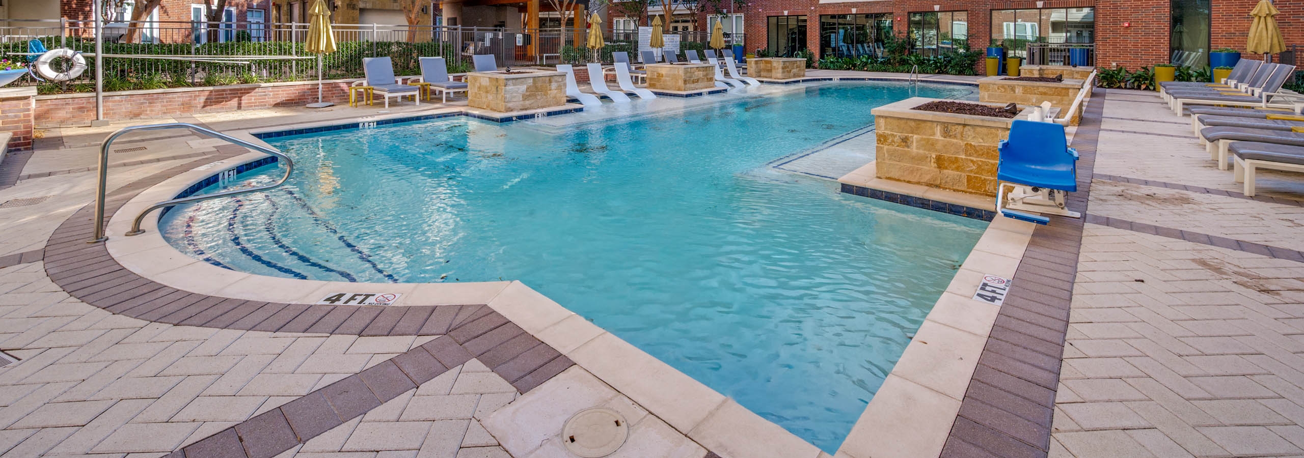 Resort-style swimming pool with white in-water loungers, umbrellas, cushioned seating and brick buildings at AMLI Frisco Crossing apartments.