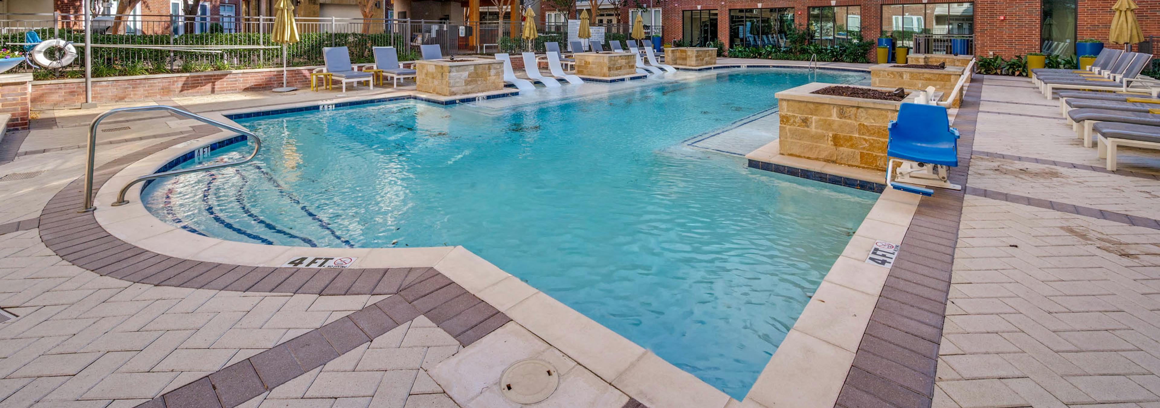 Resort-style swimming pool with white in-water loungers, umbrellas, cushioned seating and brick buildings at AMLI Frisco Crossing apartments.