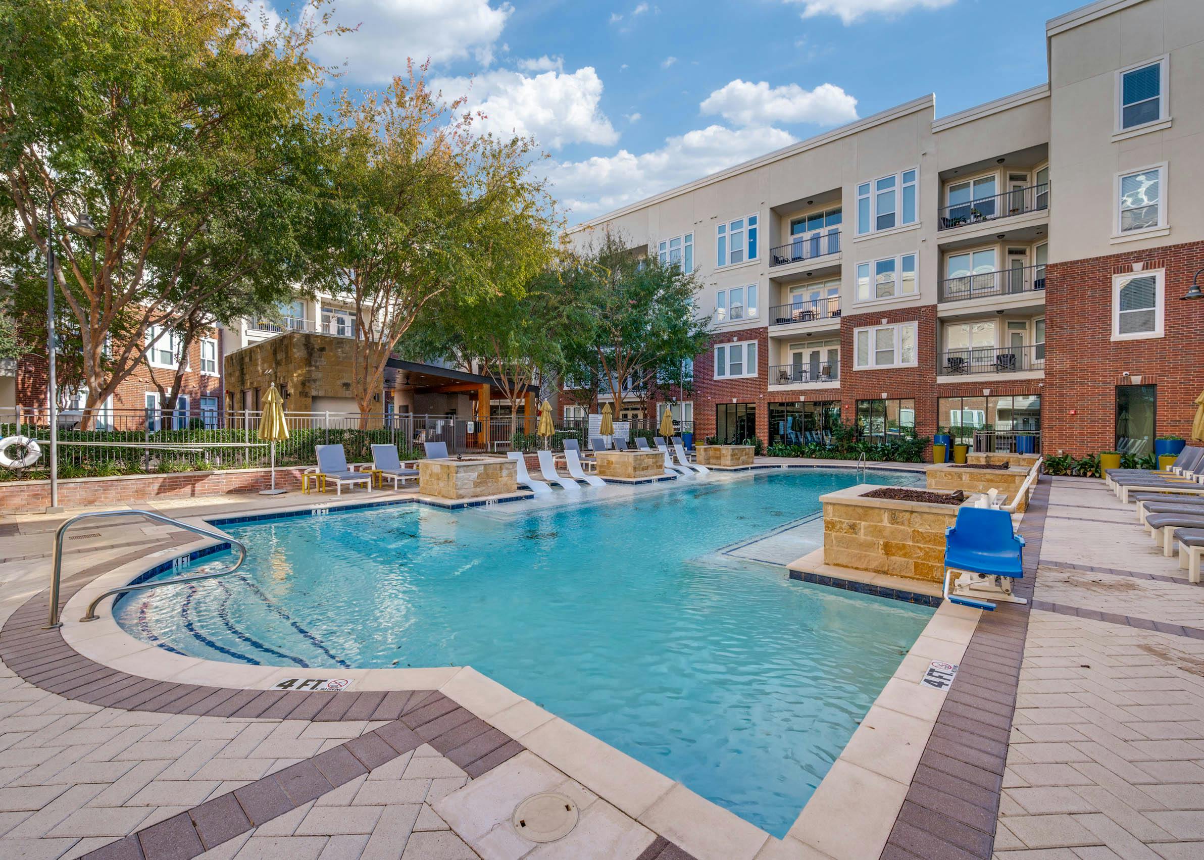 Resort-style swimming pool with white in-water loungers, umbrellas, cushioned seating and brick buildings at AMLI Frisco Crossing apartments.