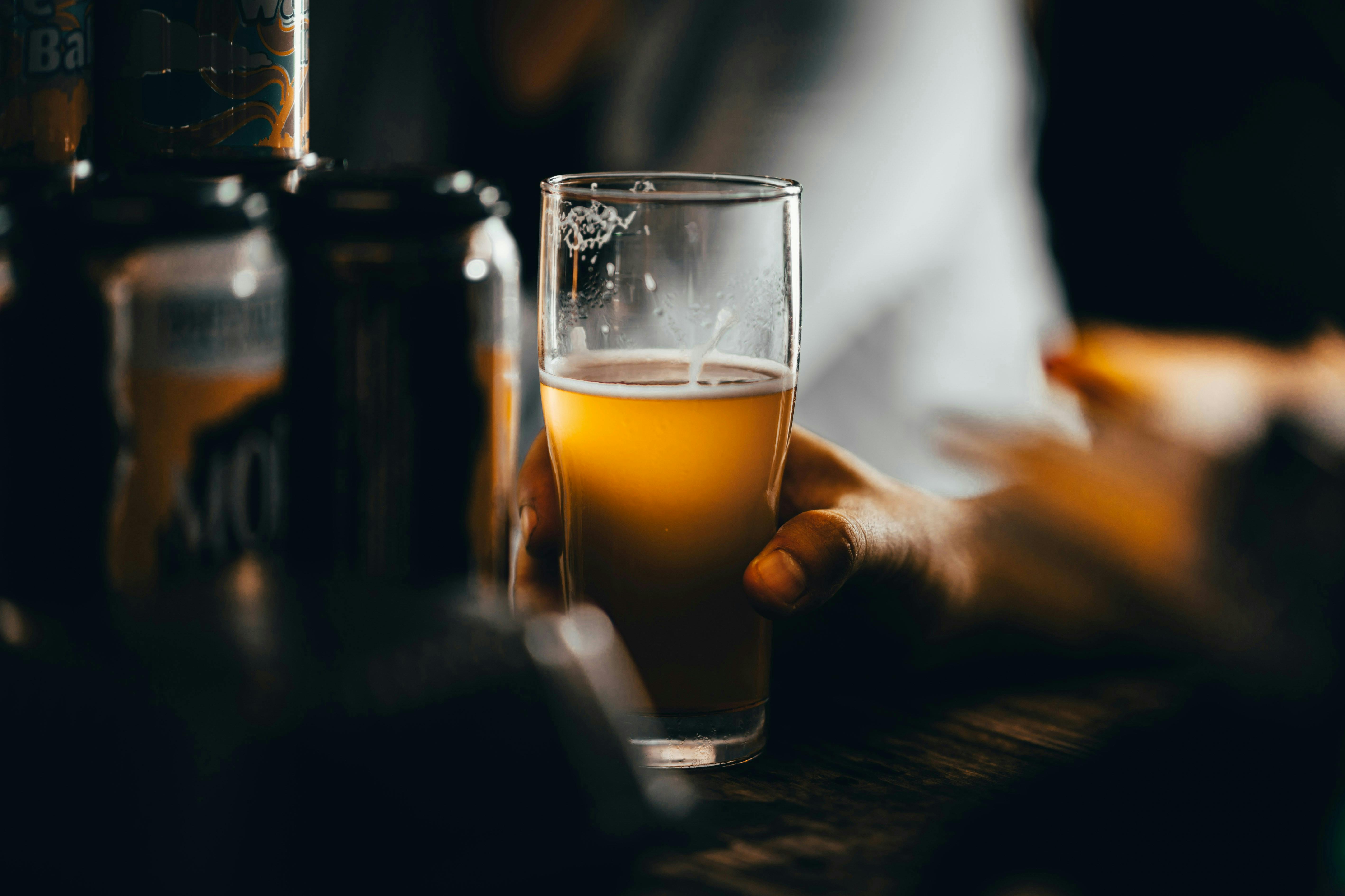 A moody, low-light shot of a hand holding a glass of hazy golden beer at a dark bar. To the left, several beer cans are stacked in the shadows, and the lighting highlights the foam and condensation on the glass.