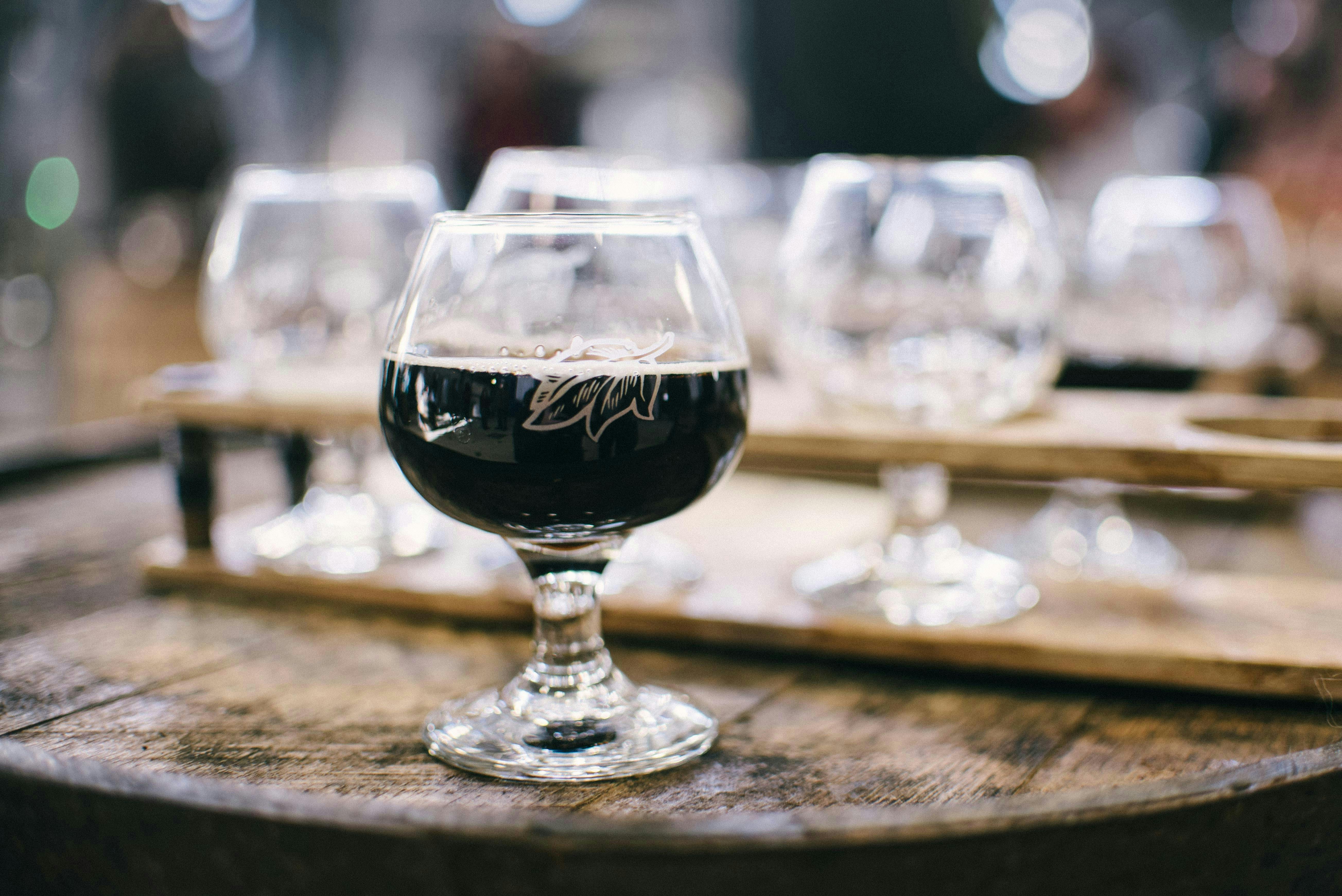 A close-up of a snifter glass filled with a very dark, opaque beer topped with a thin tan head. The glass sits on the wooden top of a barrel, with a wooden flight paddle holding empty glasses blurred in the background.