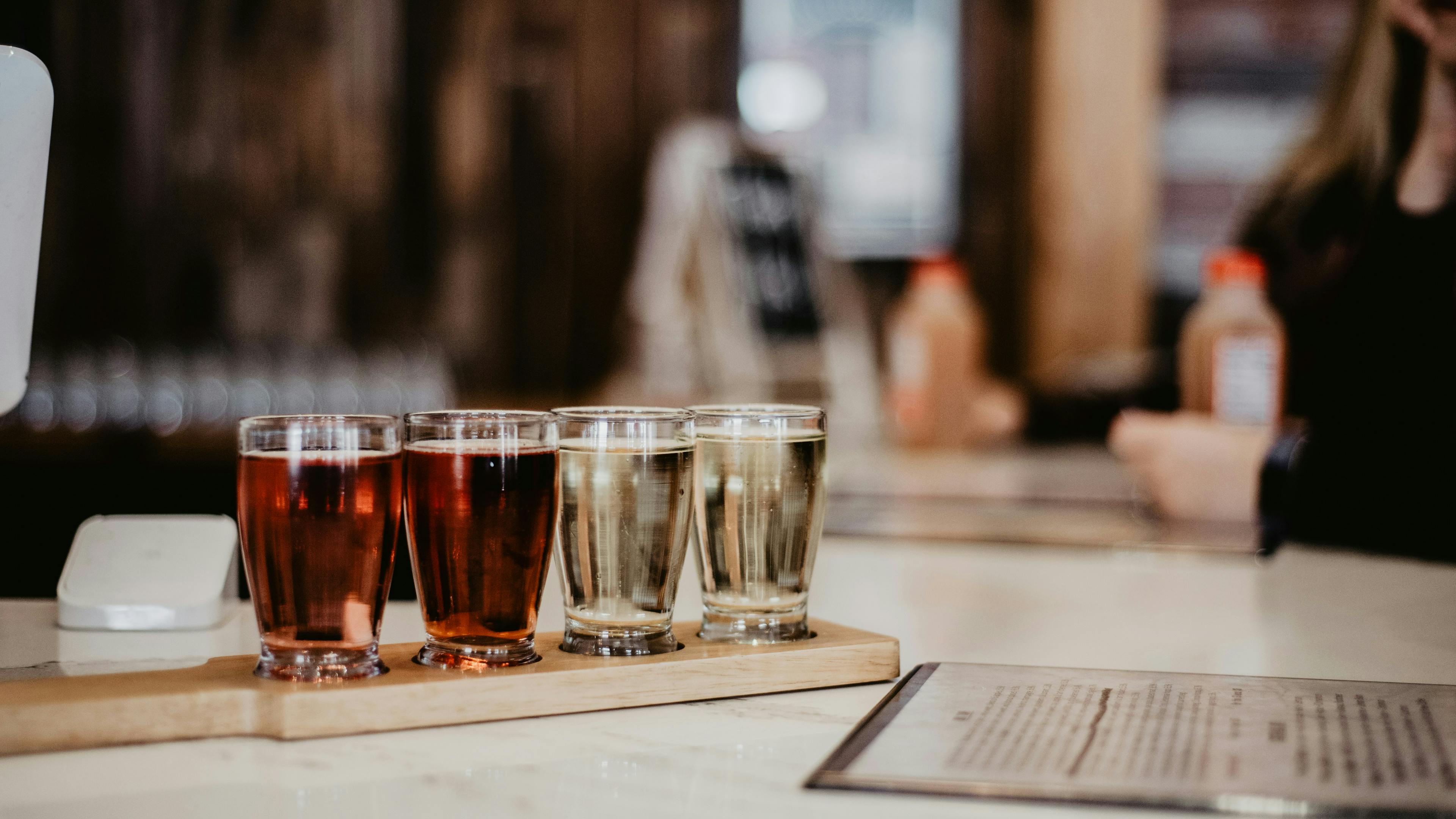 A wooden taster paddle holding four small glasses of beer on a white bar top. The flight includes two reddish-amber beers and two pale gold, clear beers, with the blurred interior of a taproom in the background.
