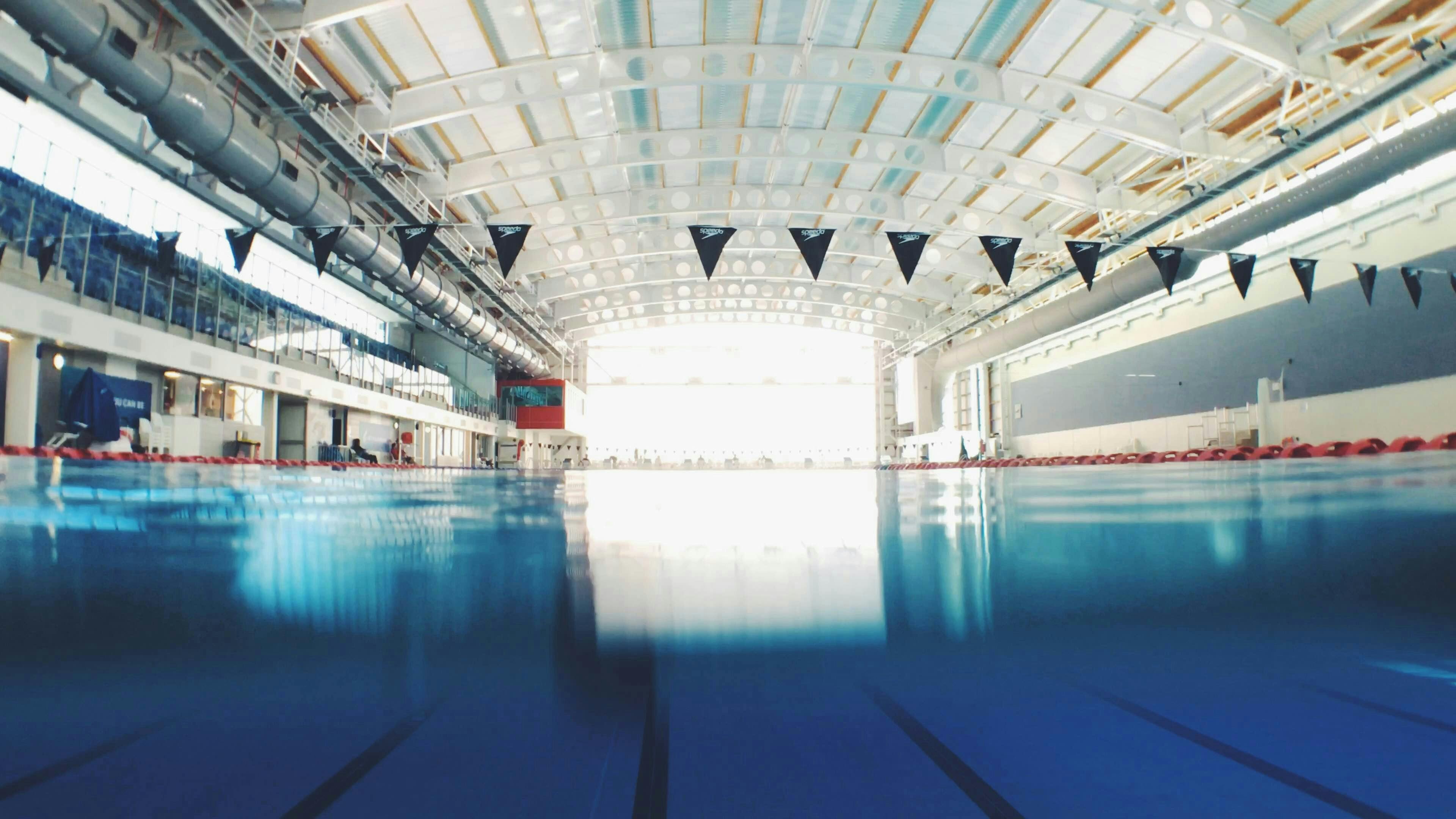A low-angle, water-level view of a large, indoor Olympic-sized swimming pool. The water is clear blue, with lane lines stretching toward the far end under a high, industrial-style arched ceiling with bright overhead lighting.
