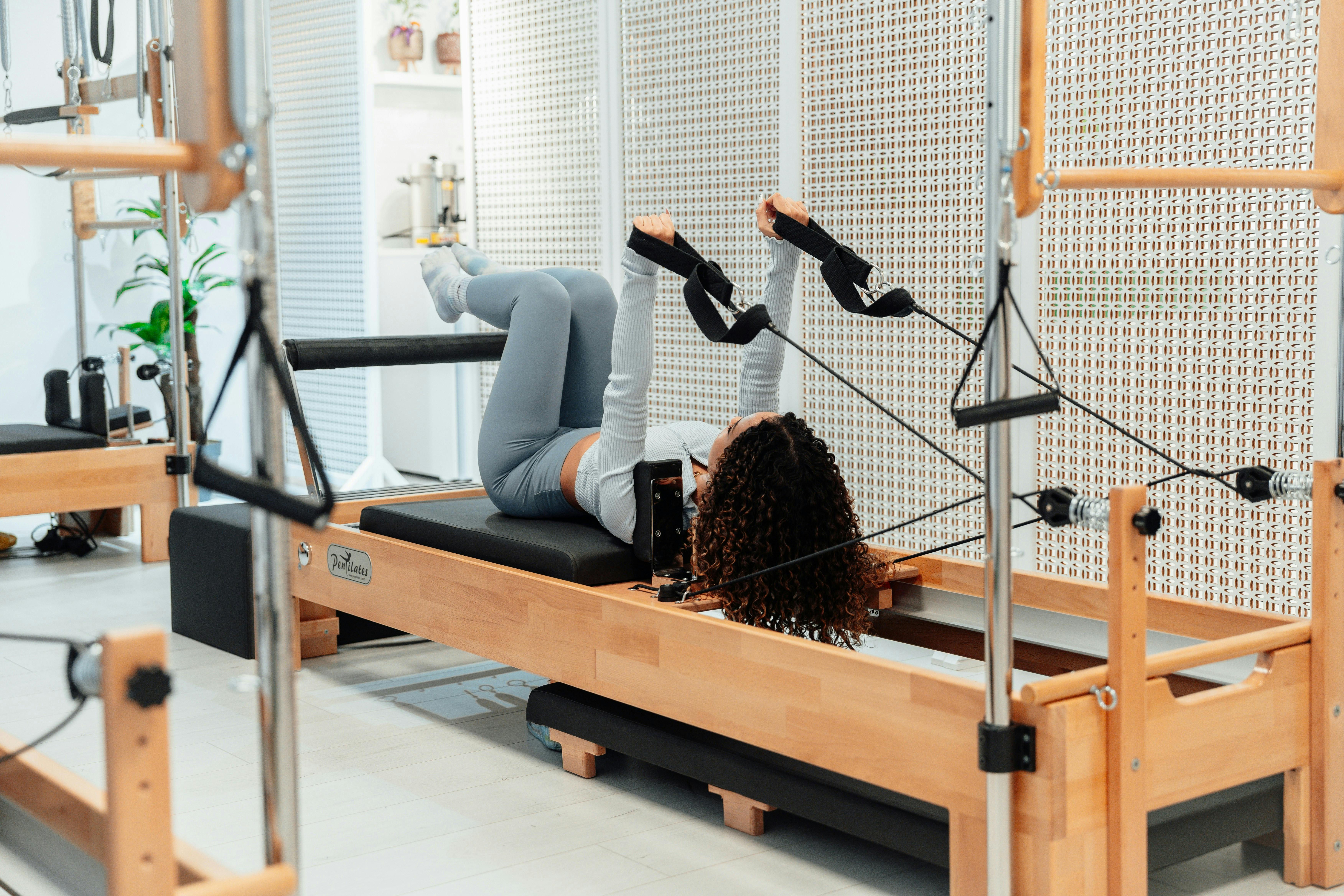 A woman lies on a wooden Pilates reformer machine, using hand straps to perform a leg and core exercise. The studio is bright and clean, featuring minimalist white decor and additional reformer equipment in the background.