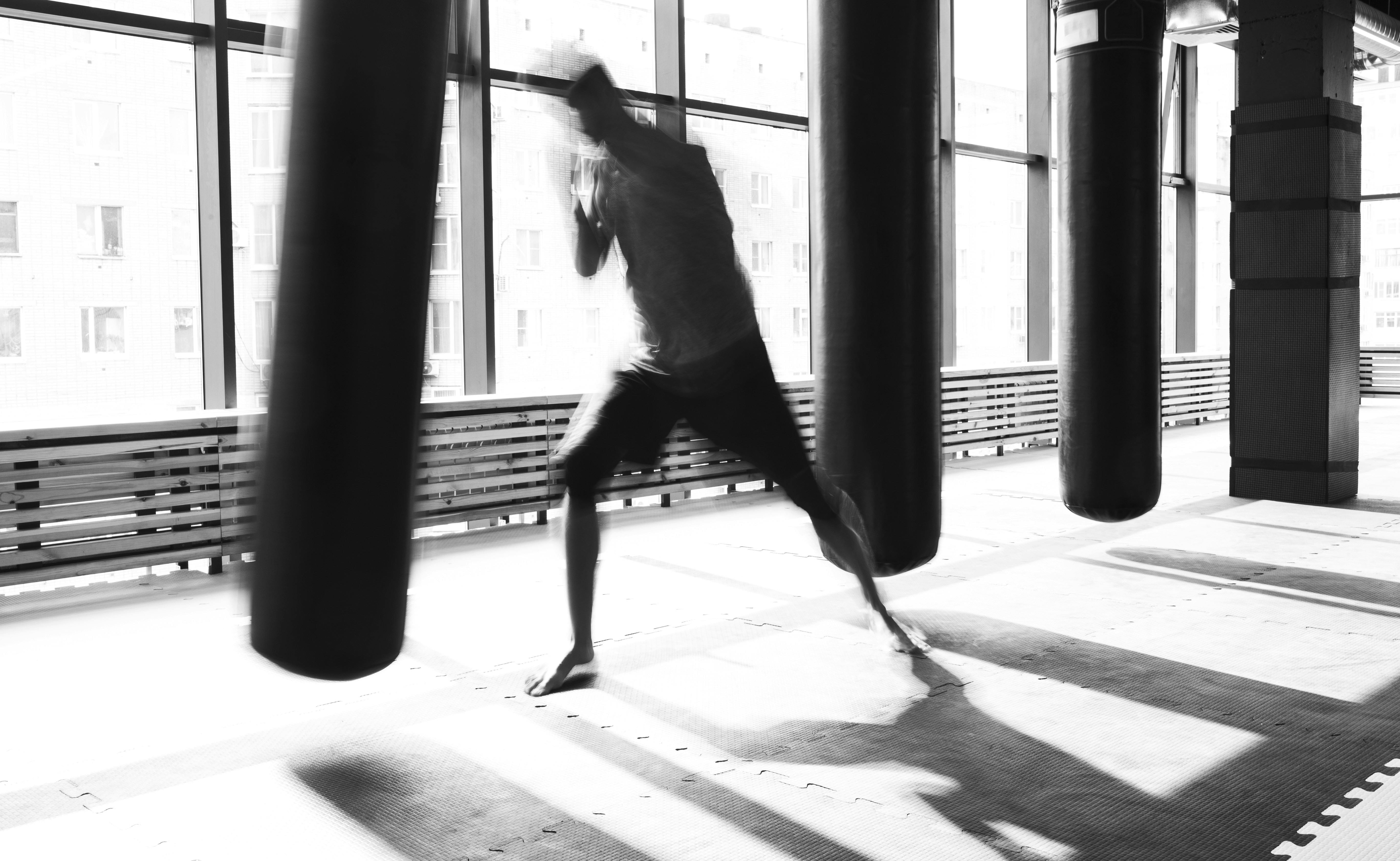 A black and white, motion-blurred action shot of a person shadowboxing between heavy punching bags. The room is bright with natural light from large windows in the background, and the floor is covered in protective foam mats.