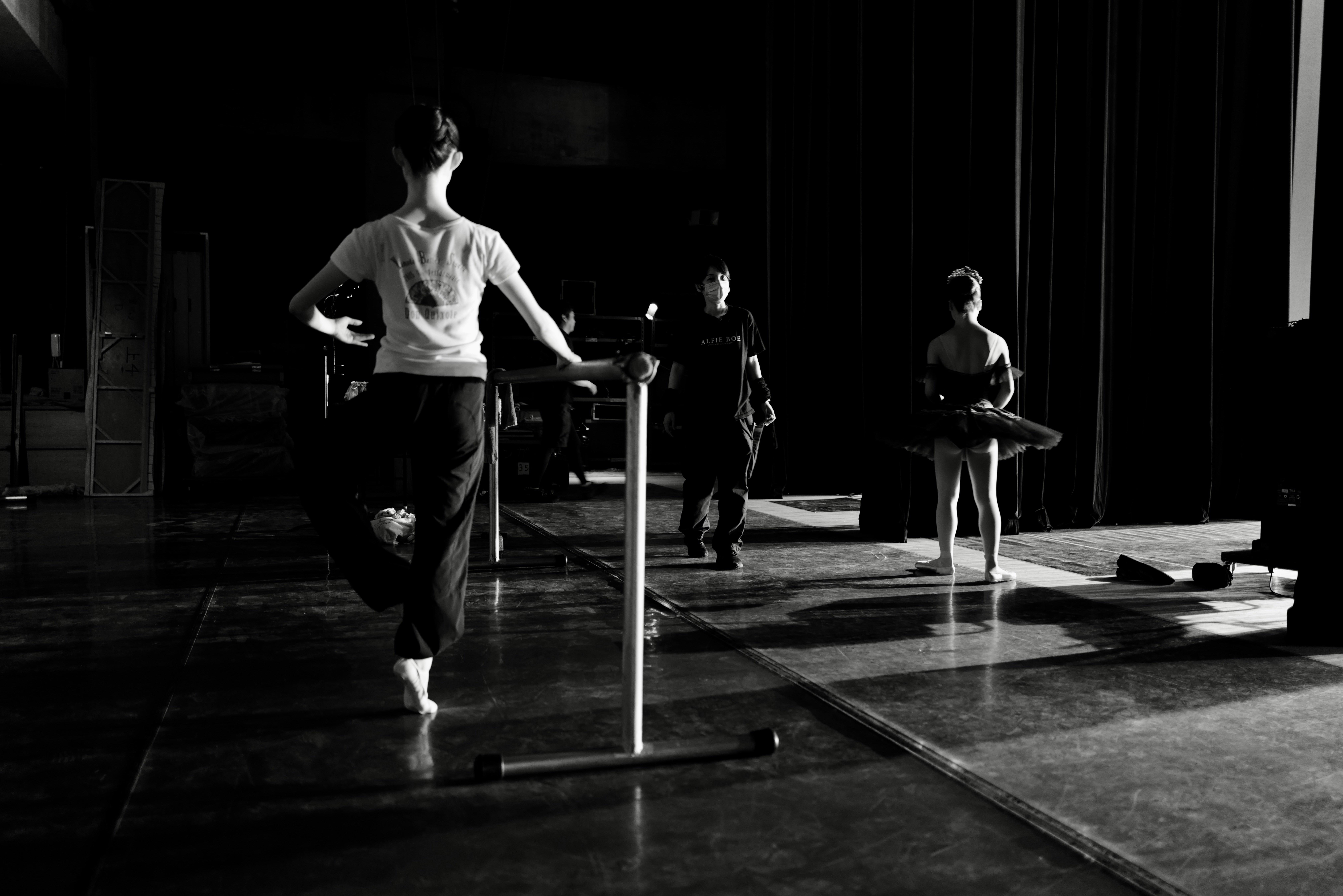 A black and white photo from behind a dancer at a ballet barre in a dimly lit studio. Another dancer in a tutu stands further back, and a masked instructor stands in the center of the dark wooden floor.