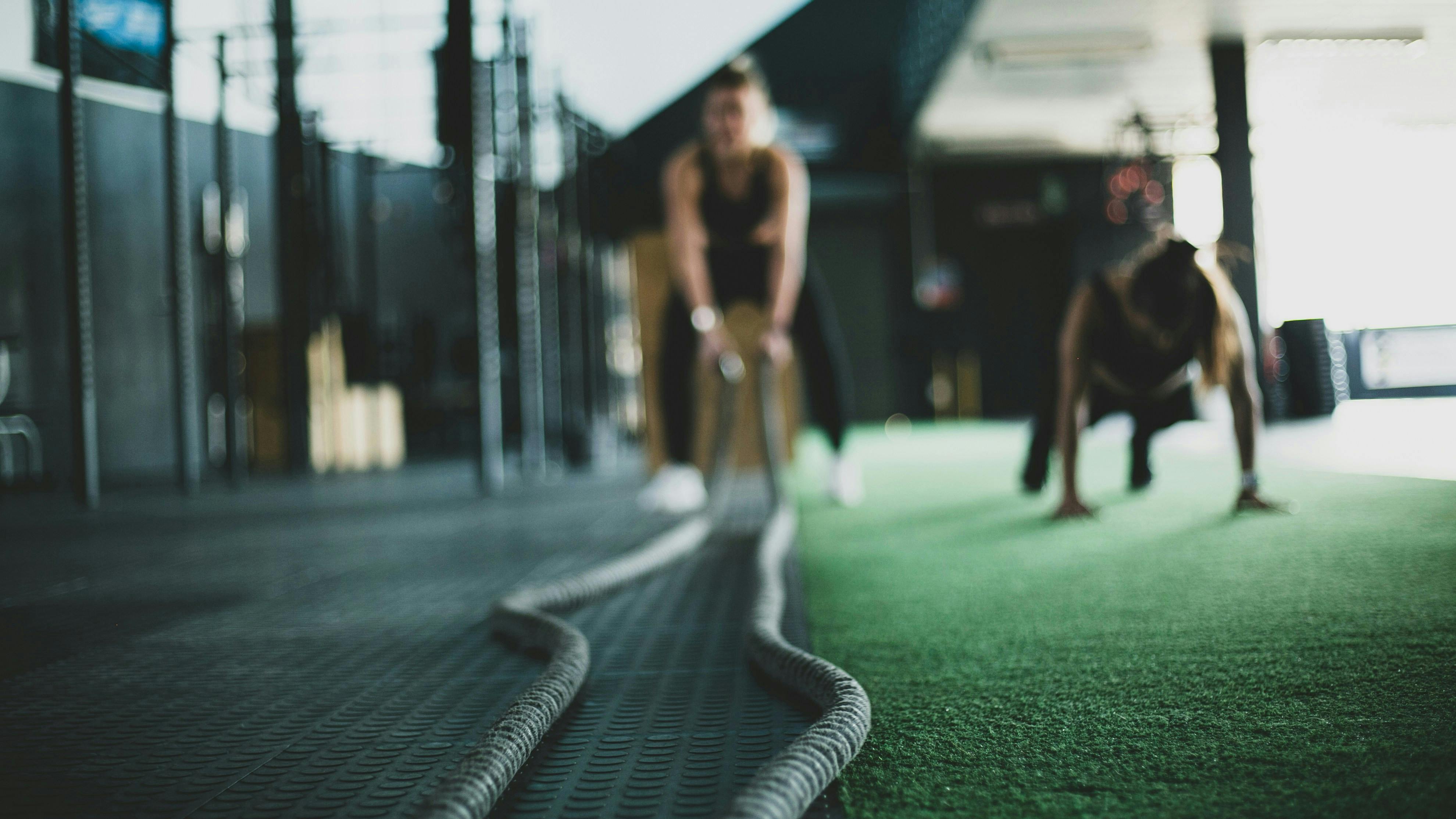 A shallow depth-of-field shot focusing on heavy battle ropes snaking across a black gym floor. In the blurred background, a person is preparing to use the ropes on a green turf section of a functional fitness gym.