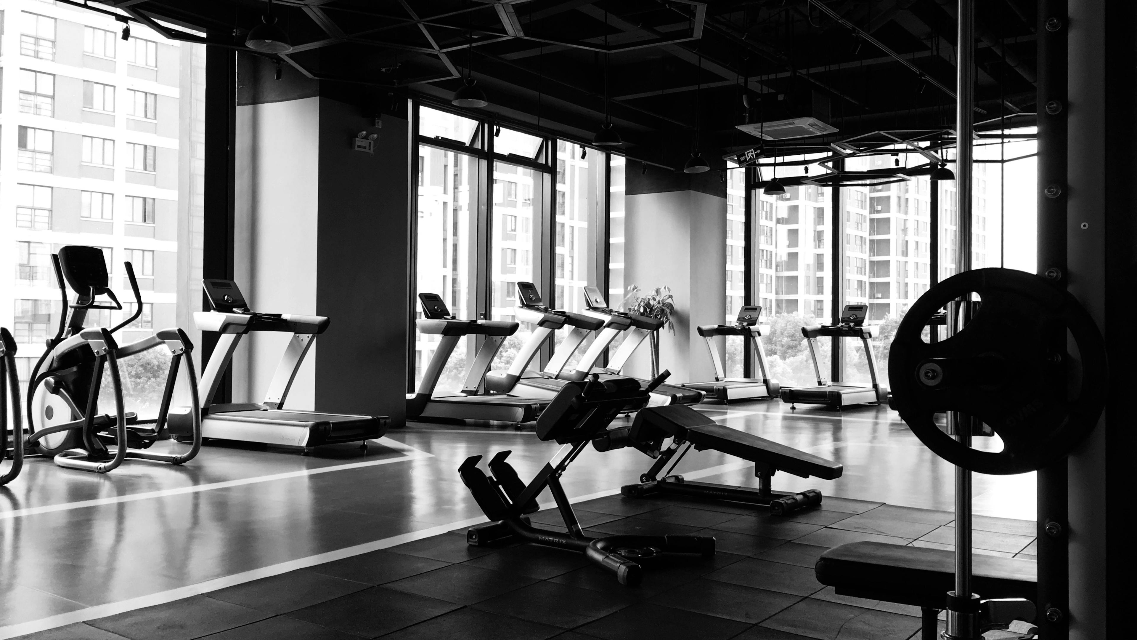A black and white, high-angle shot of a modern gym. A row of treadmills and elliptical machines faces large floor-to-ceiling windows overlooking city buildings. In the foreground, weight benches and a Smith machine are positioned on dark rubber flooring.