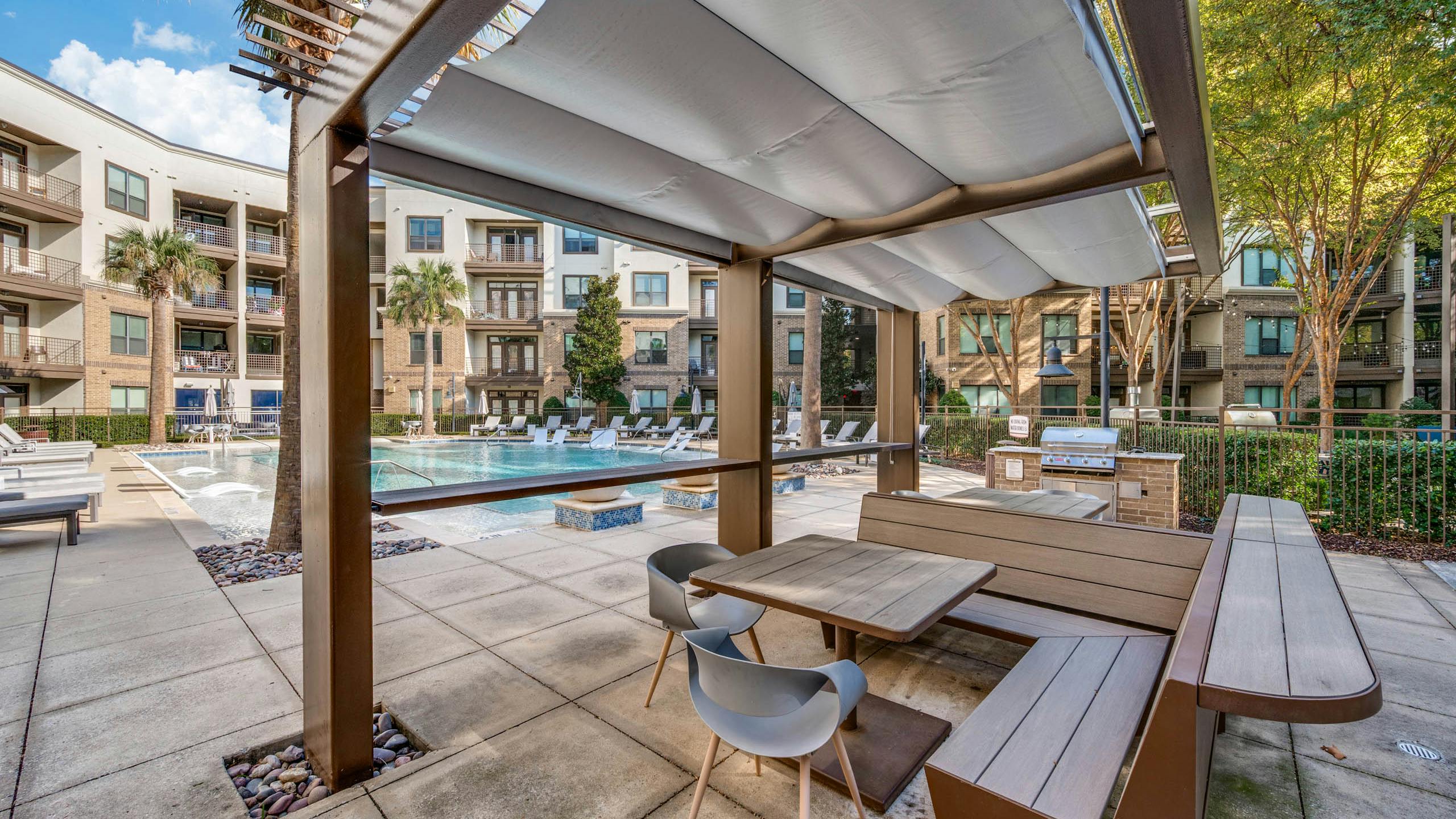 Poolside view at AMLI at the Ballpark apartments with lounge chairs, umbrellas, and landscaped trees surrounding clear blue water.