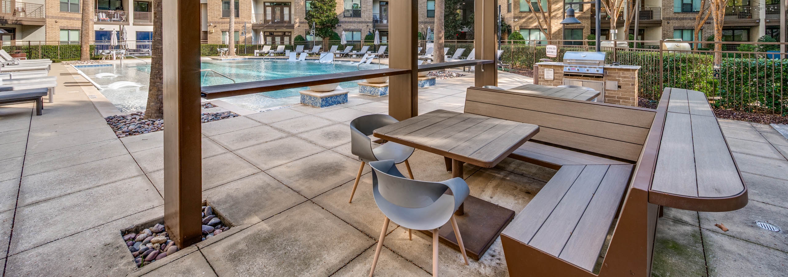 Poolside view at AMLI at the Ballpark apartments with lounge chairs, umbrellas, and landscaped trees surrounding clear blue water.