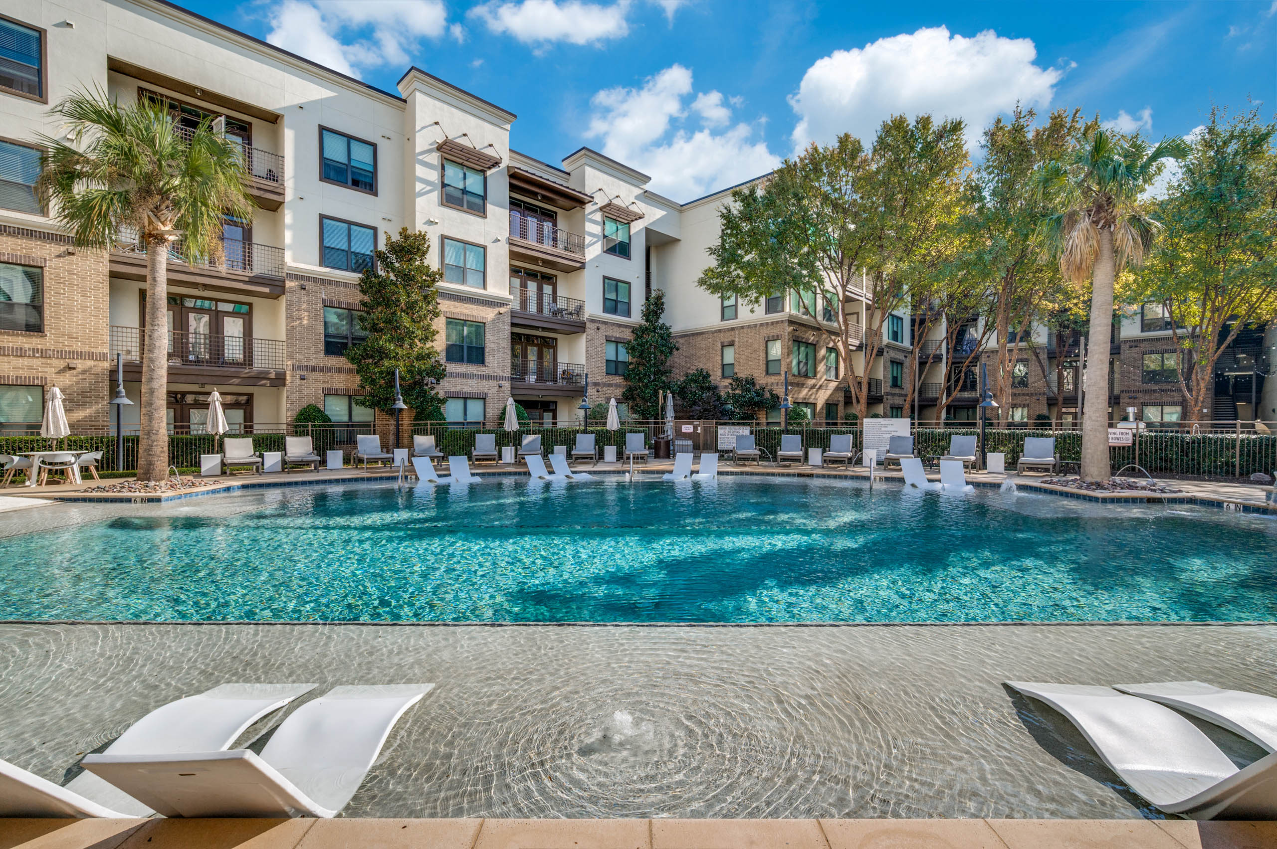Poolside view at AMLI at the Ballpark apartments with lounge chairs, umbrellas, and landscaped trees surrounding clear blue water.