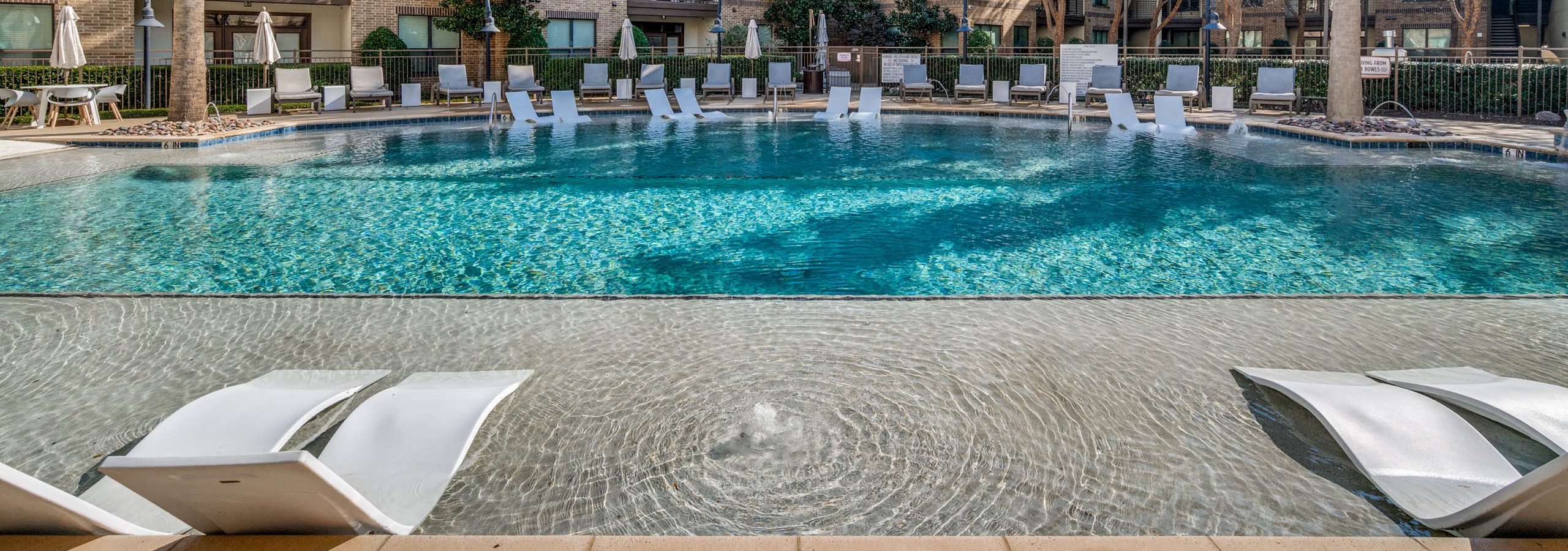 Poolside view at AMLI at the Ballpark apartments with lounge chairs, umbrellas, and landscaped trees surrounding clear blue water.
