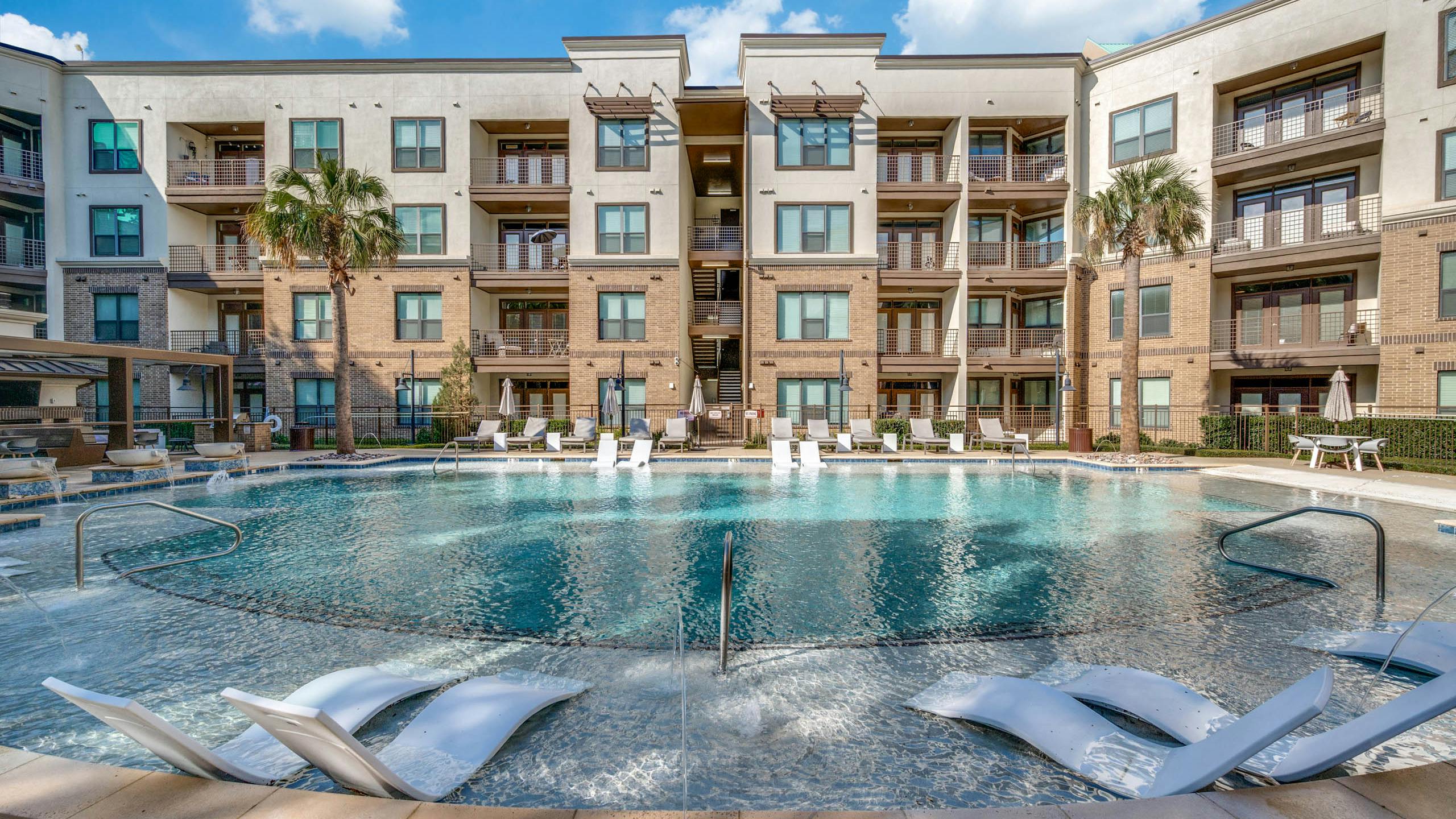 Straight-on view of AMLI at the Ballpark apartments with large pool, sun loungers in shallow area, and palm trees under a blue sky.