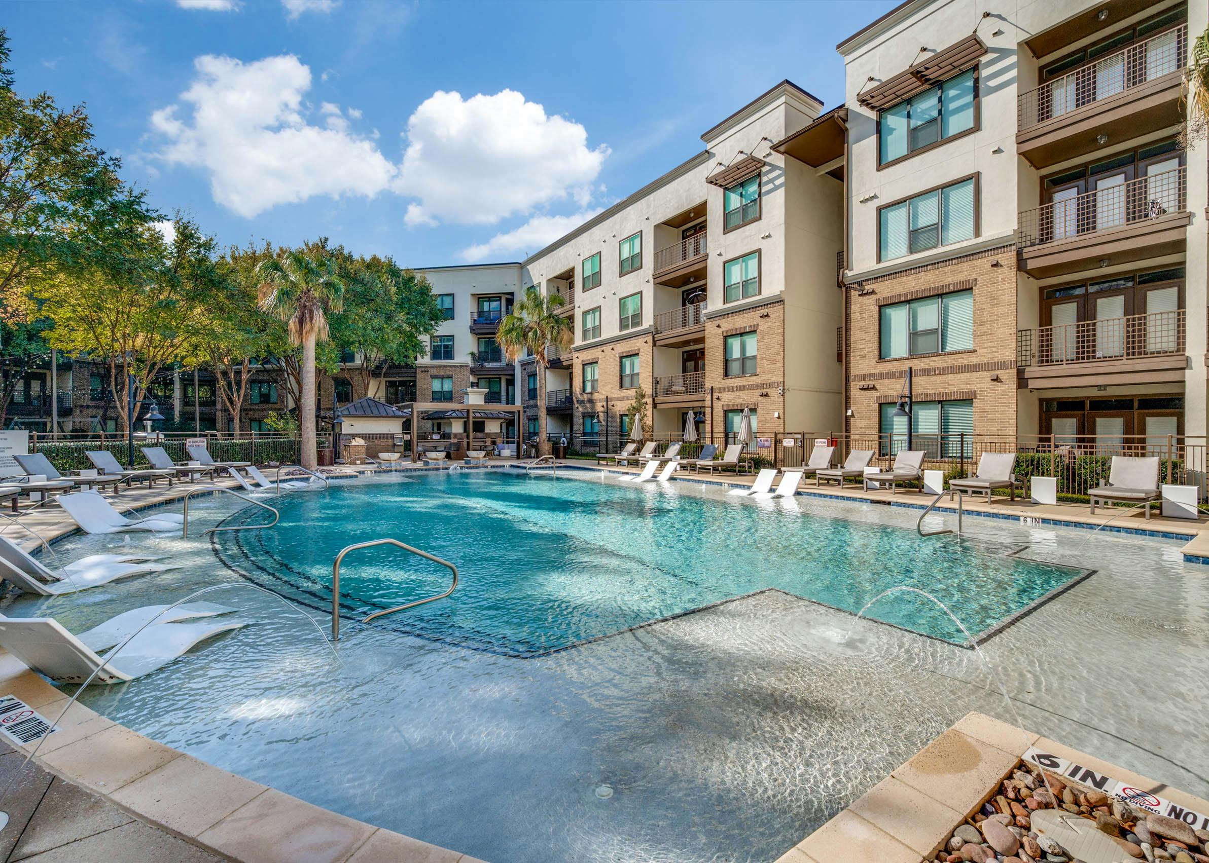 Wide bright view of the resort-style pool with in-water loungers, palm trees, and sun deck at AMLI at the Ballpark apartments.