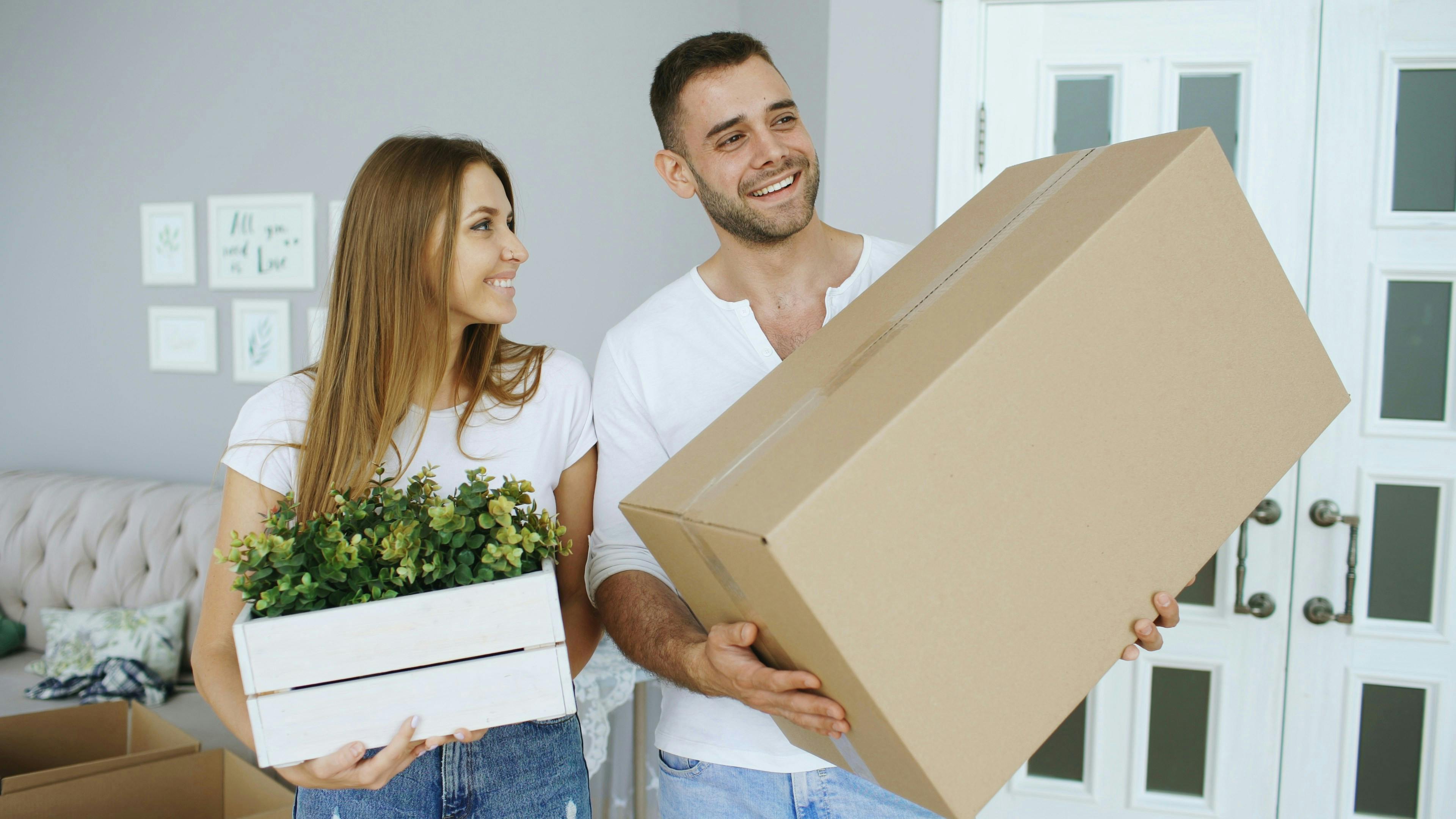 A couple with moving boxes and a houseplant standing beside each other in a brightly lit room