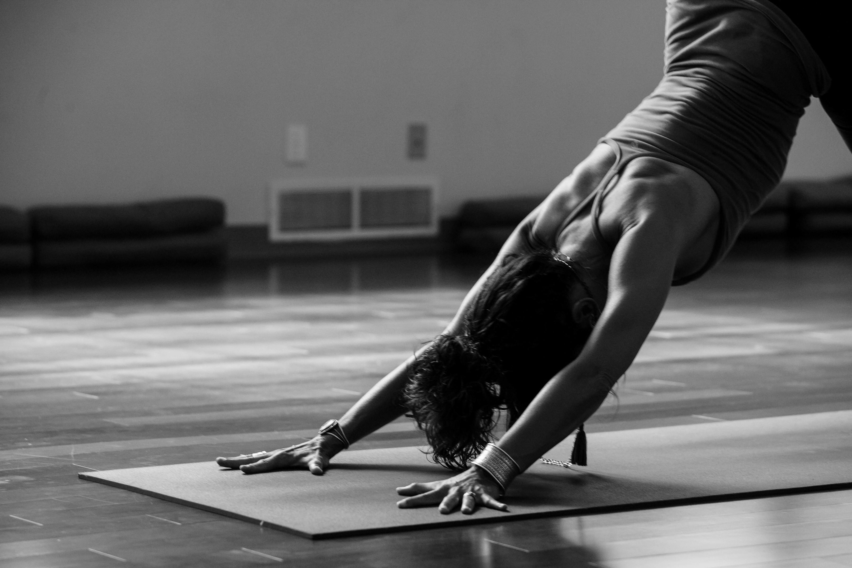 A black and white, side-profile image of a person practicing yoga in a downward-facing dog pose. They are positioned on a mat in a spacious studio with polished wooden floors, emphasizing strength and flexibility.
