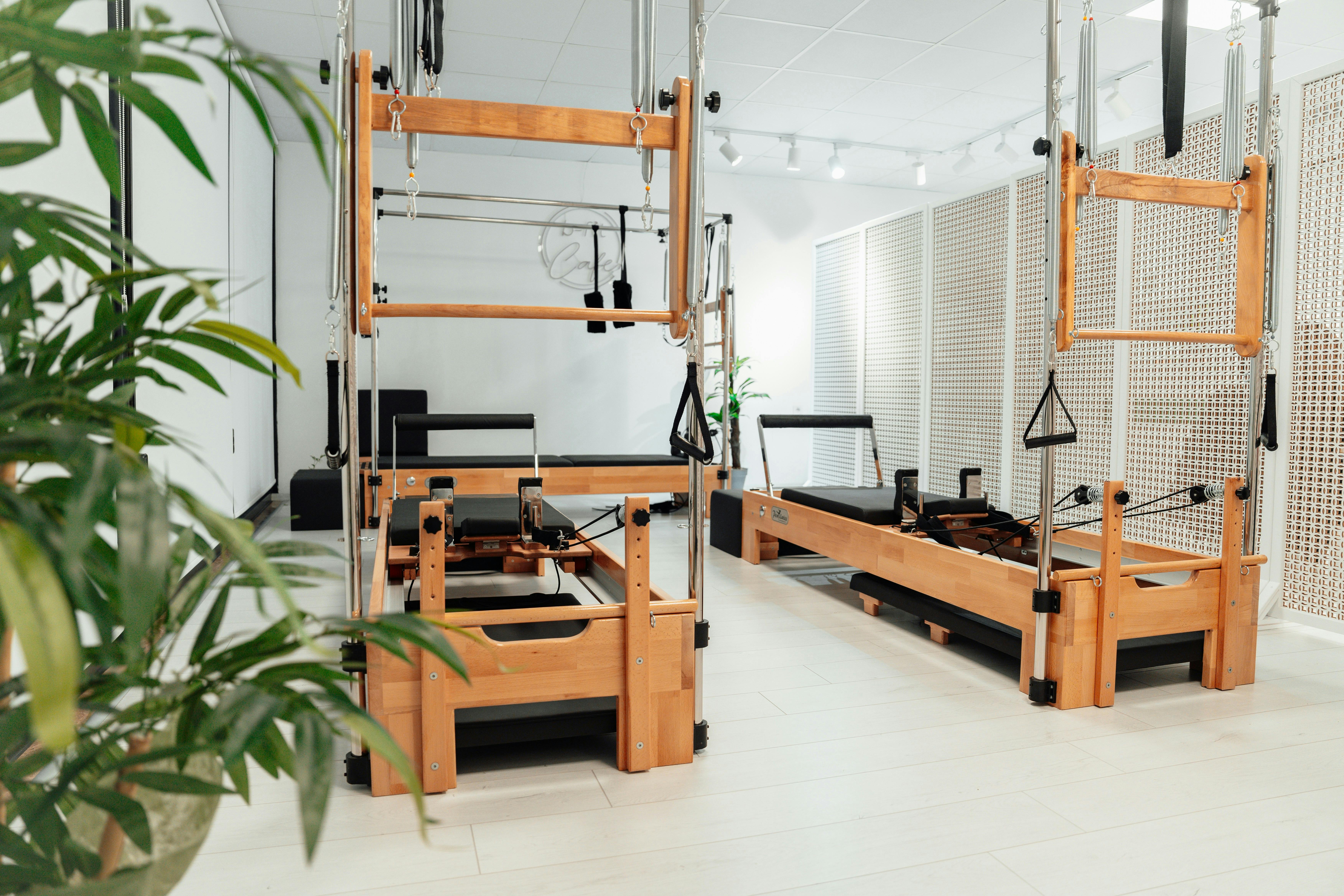 A wide, bright shot of a clean Pilates studio featuring multiple wooden reformer machines. The room is minimalist with white walls, light-colored flooring, and a green plant in the foreground, creating a serene environment for exercise.