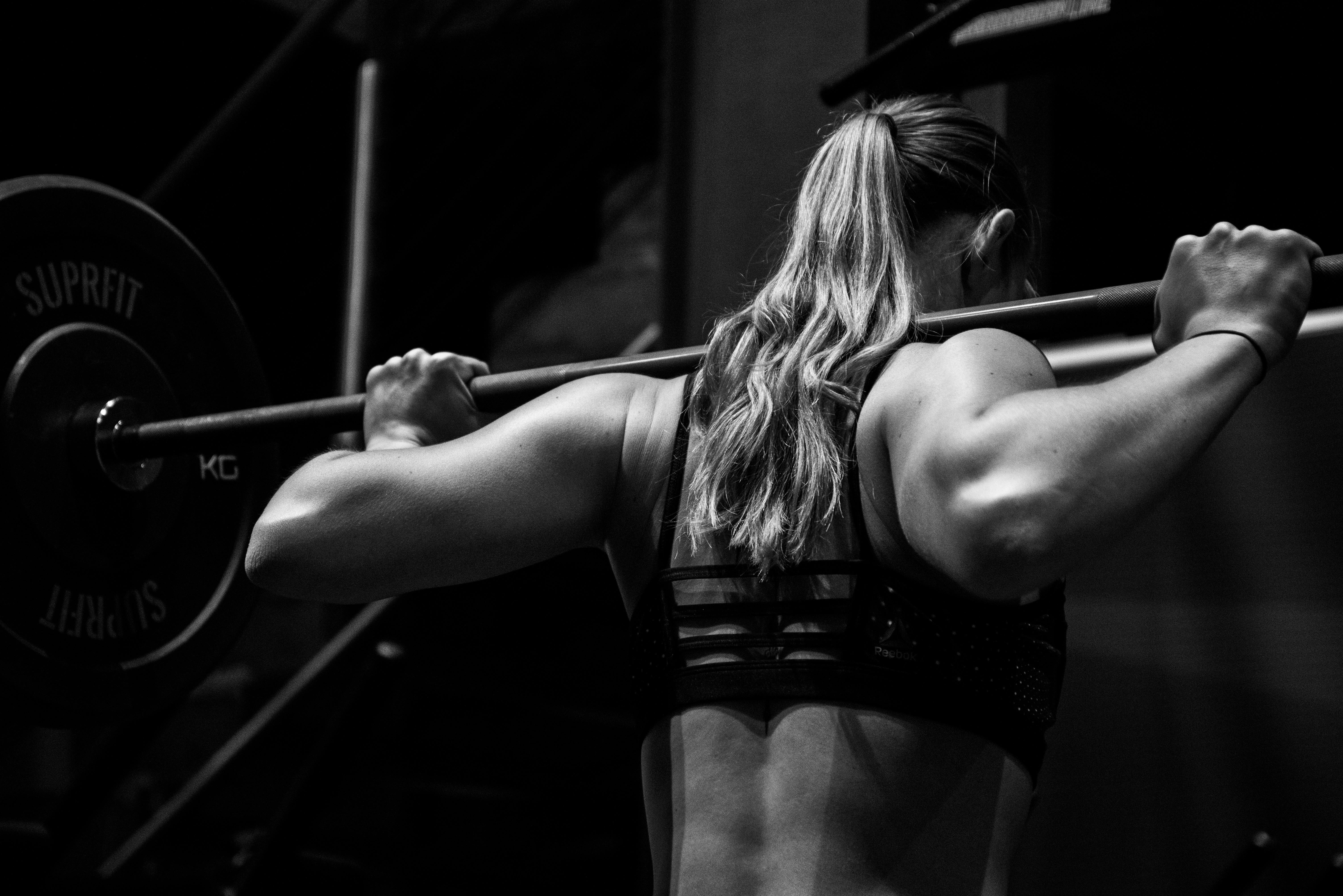 A dramatic, black and white close-up from behind a person performing a barbell squat. The focus is on the muscular definition of their back and shoulders as they grip the bar, which is loaded with heavy weights labeled "SUPRFIT."