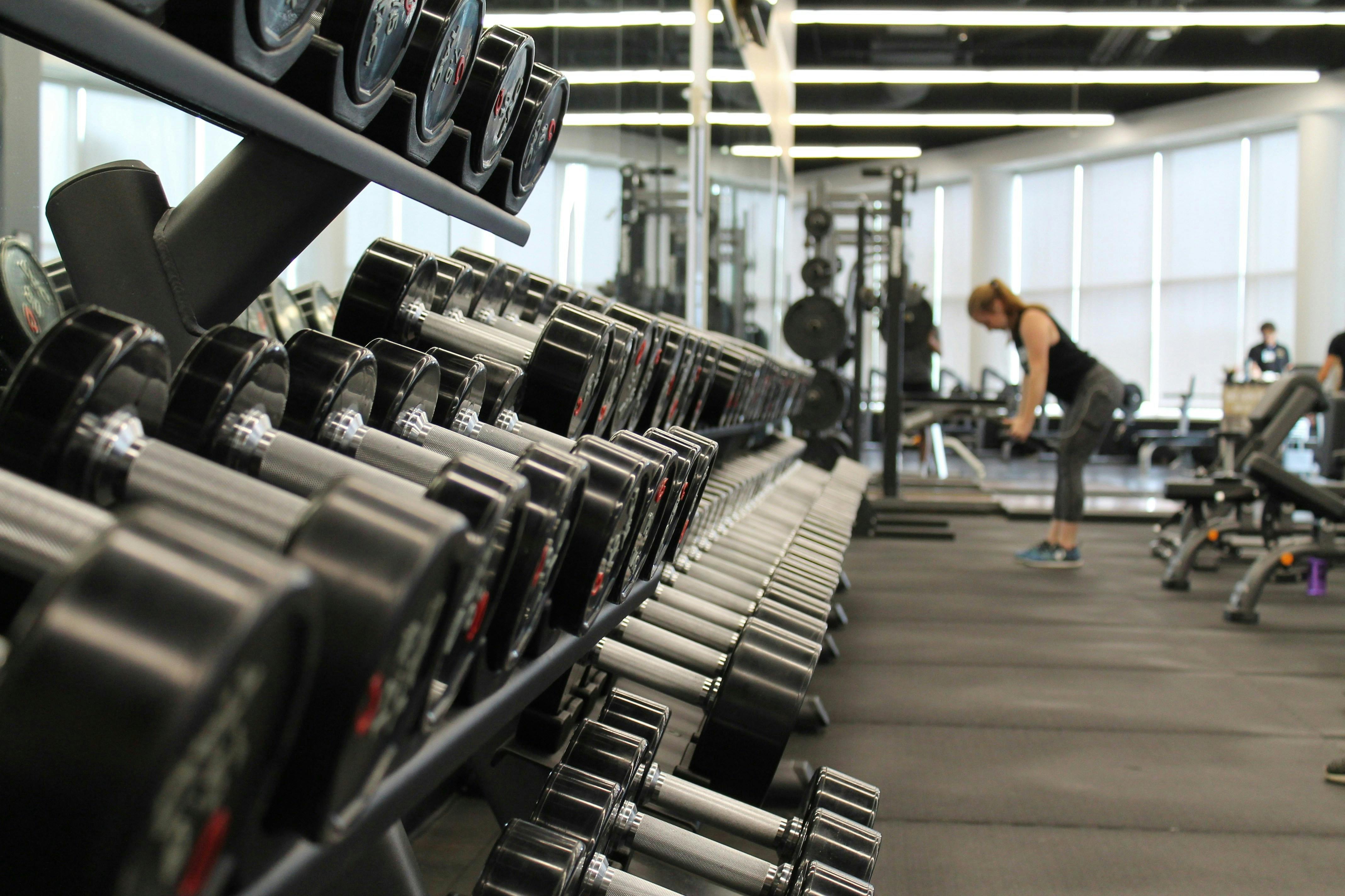 A perspective shot focusing on a long rack of identical black and silver dumbbells in a brightly lit gym. In the blurred background, a woman is seen exercising near other weightlifting equipment and large windows.