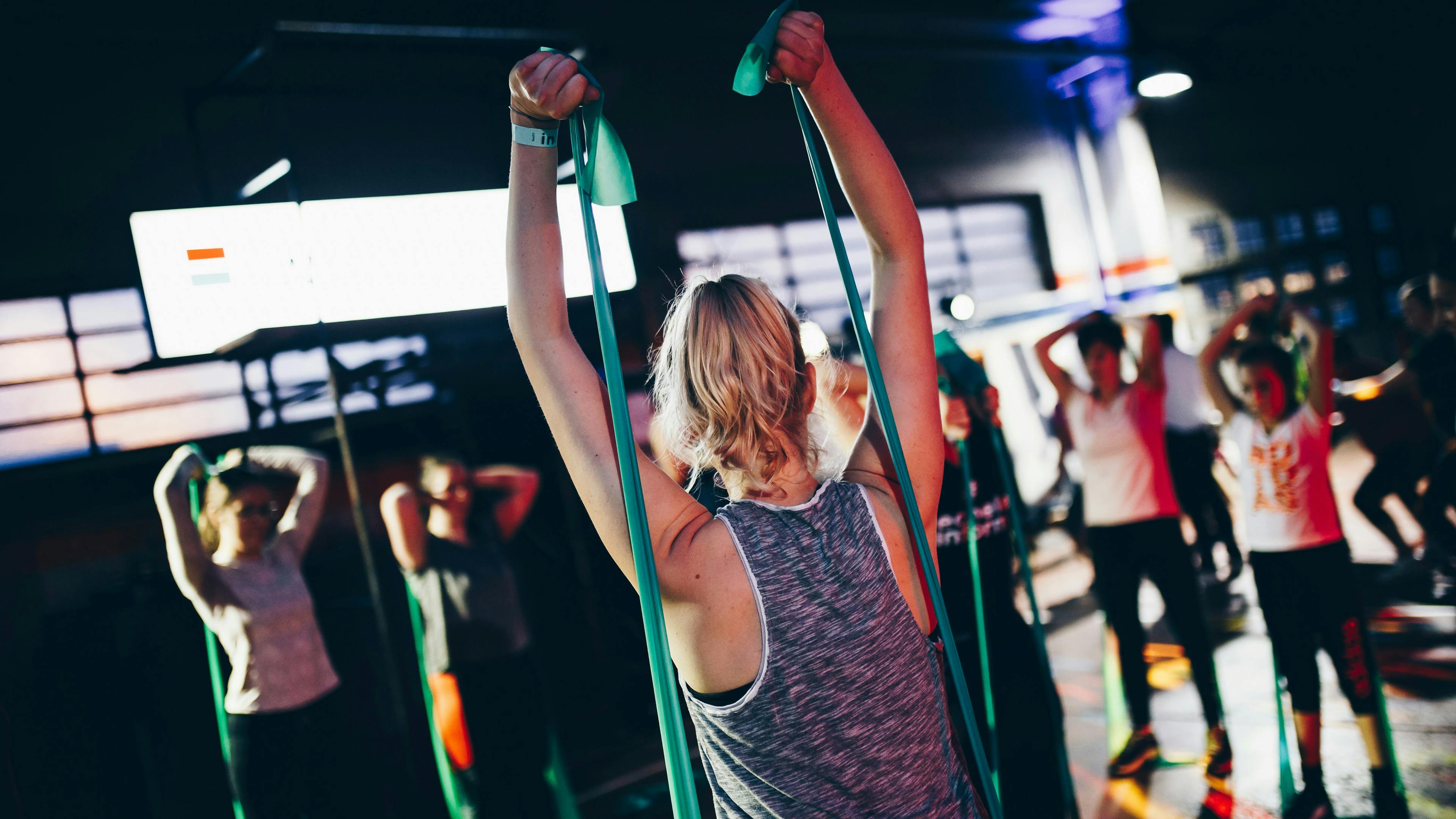 A view from behind a woman in a grey tank top during a group exercise class. She and other participants are holding green resistance bands high above their heads in a dark, energetic gym setting with ambient blue and white lighting.