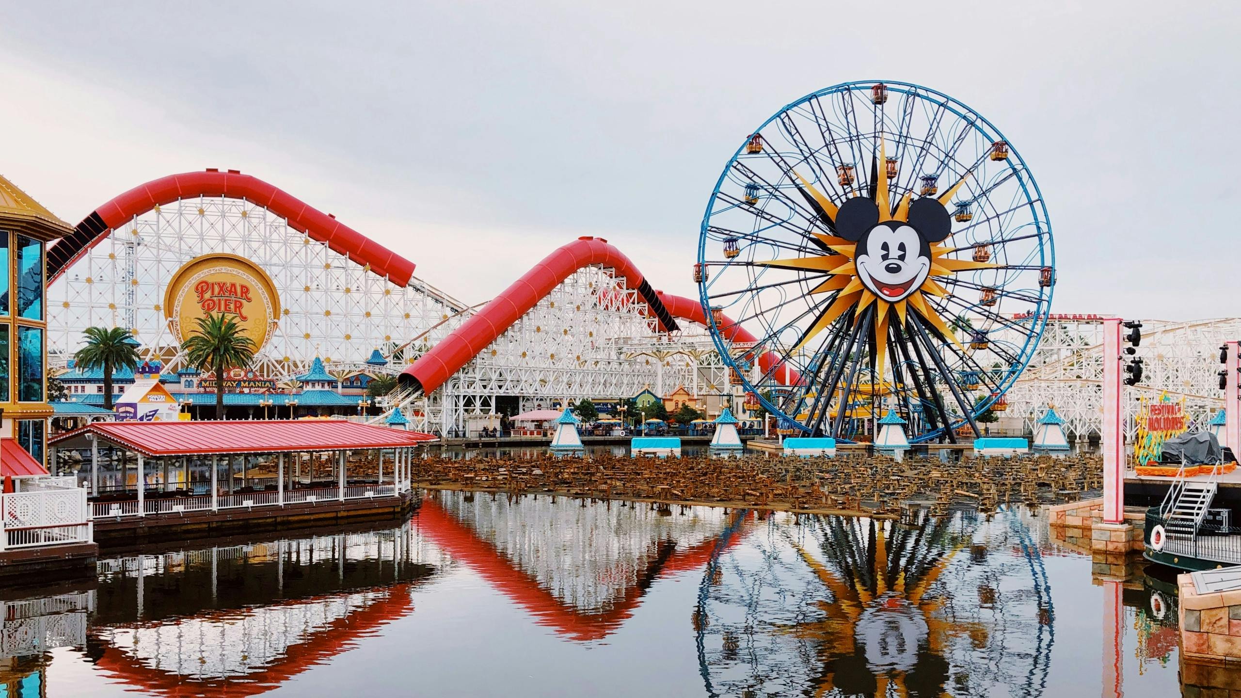 A colorful amusement park waterfront with a large Ferris wheel and red roller coaster under a bright, overcast sky.