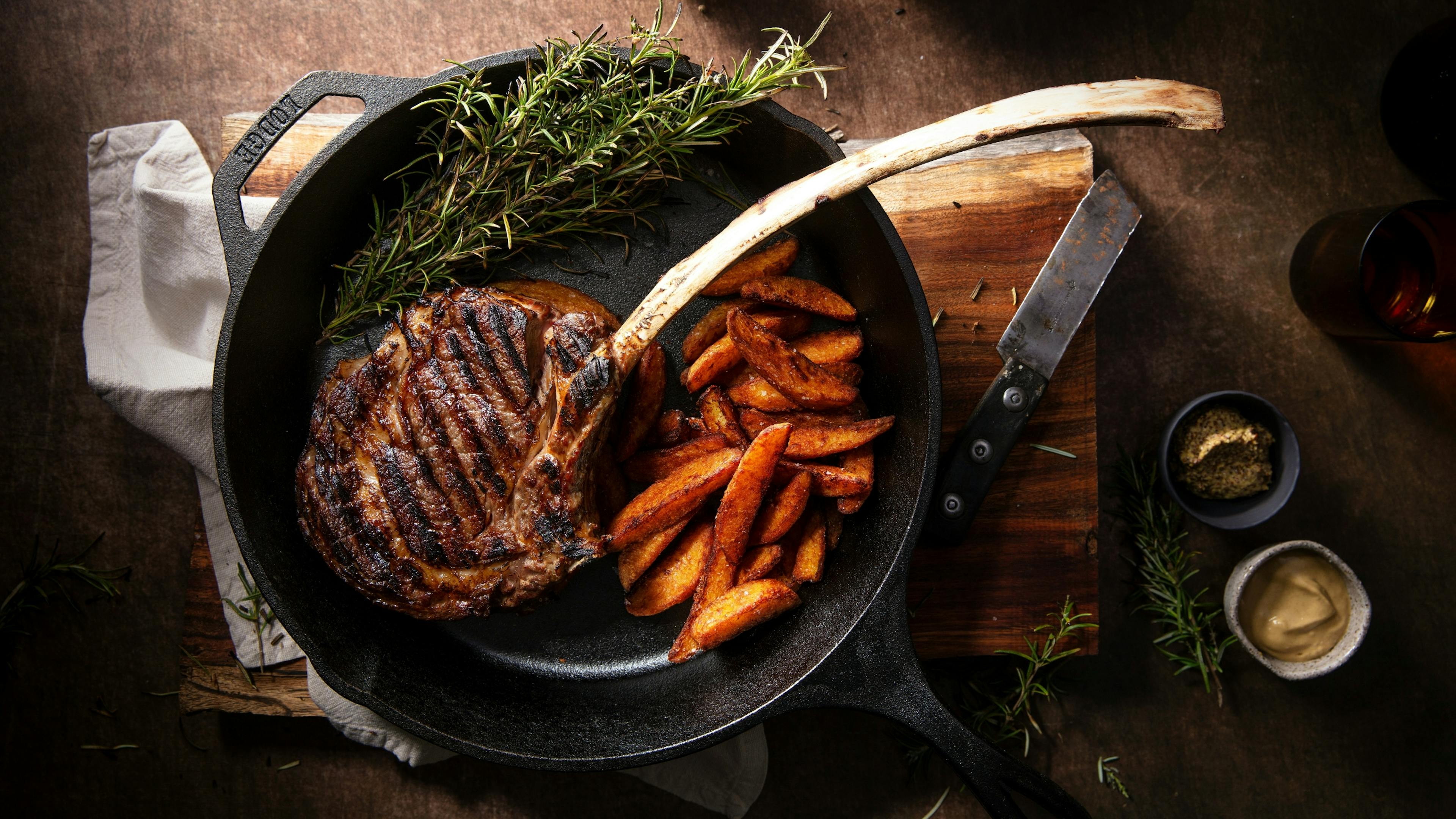 A grilled steak and potato wedges in a cast-iron skillet, garnished with fresh rosemary on a wooden board.