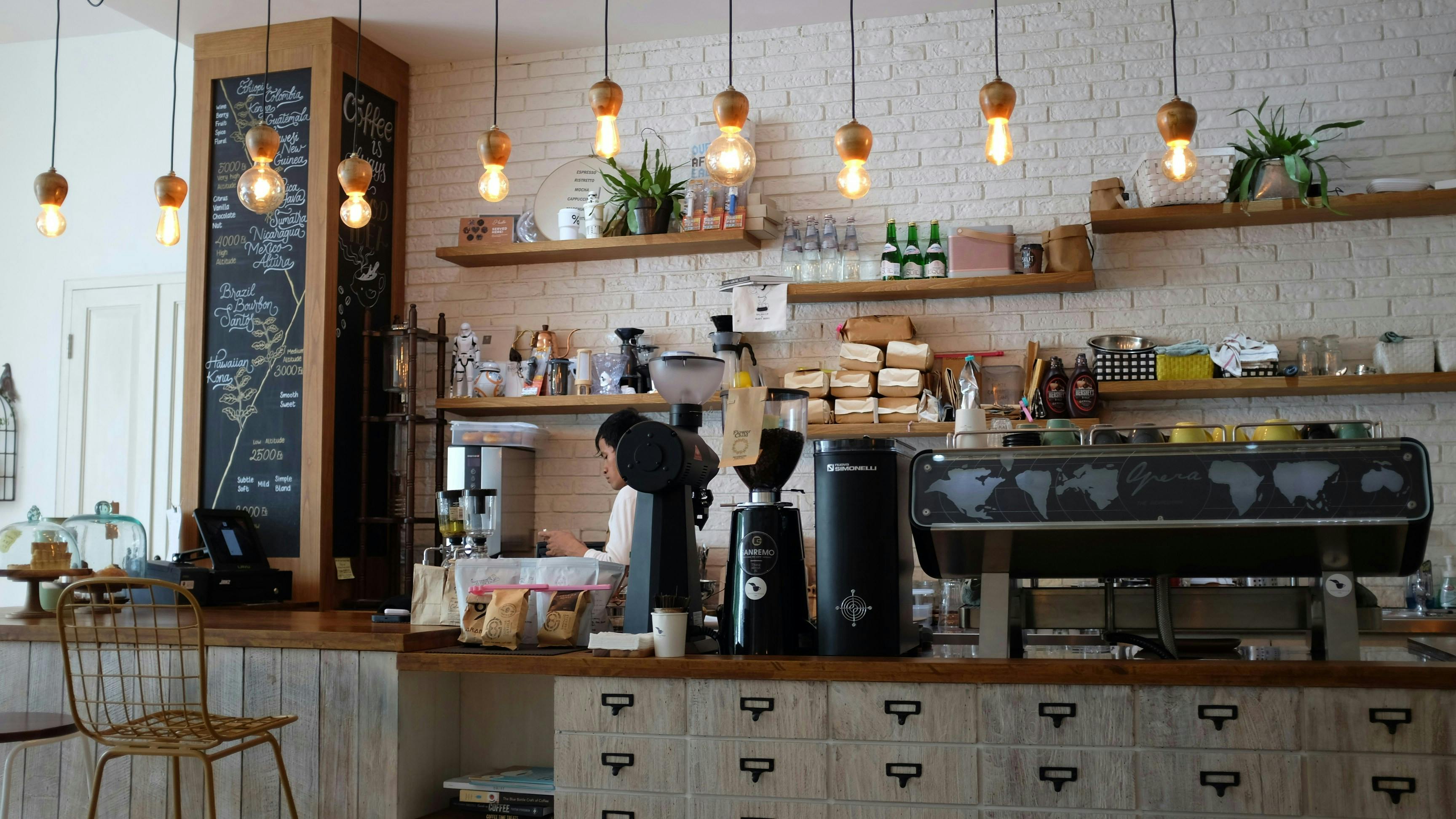 A bright, modern coffee shop counter with white brick walls, wooden floating shelves, industrial hanging Edison bulbs, and professional espresso equipment including a world-map etched coffee machine.