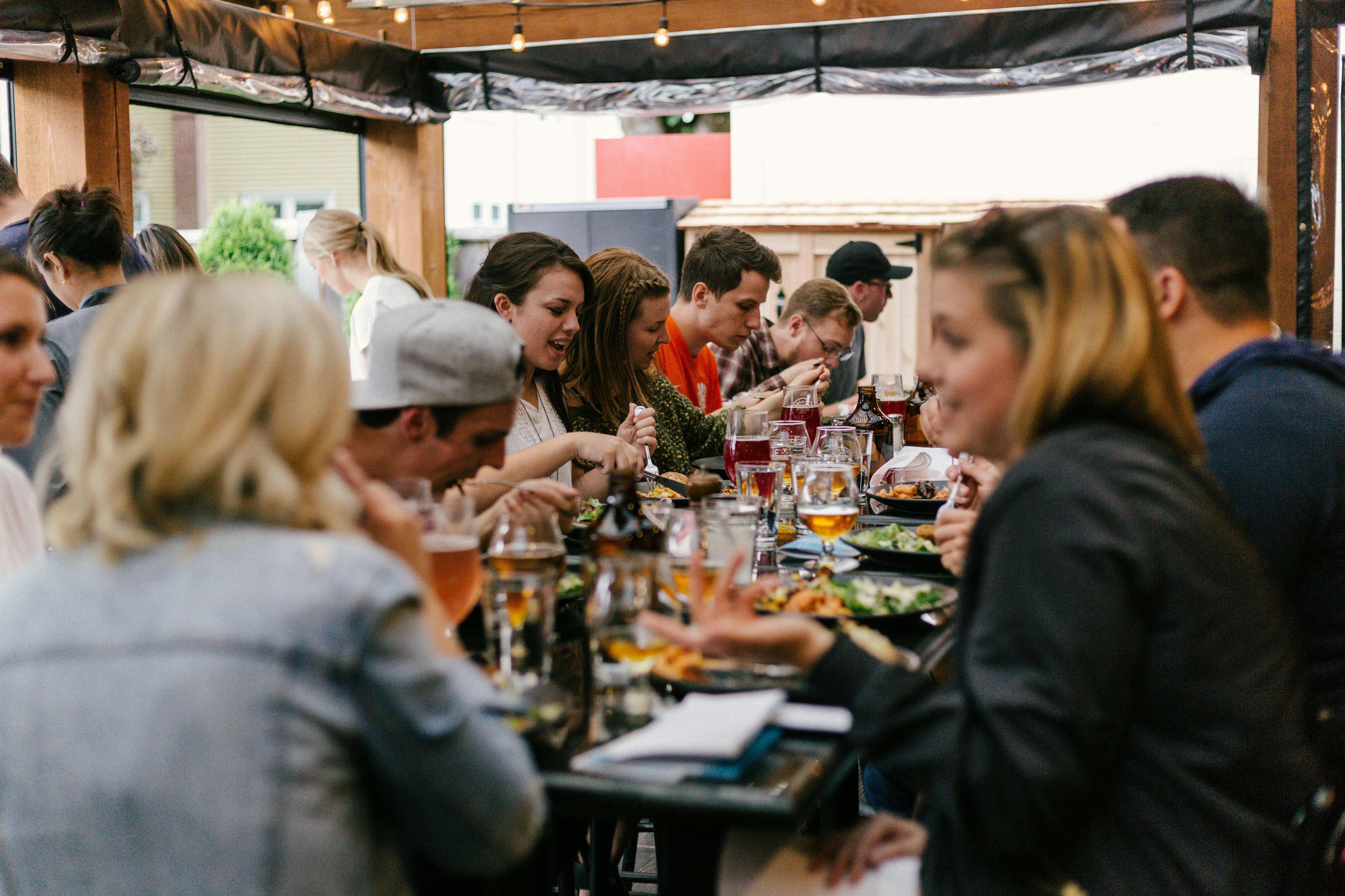 A group of friends laughing and talking while sharing a meal and glasses of beer at a long communal table in a lively, covered outdoor patio or beer garden.