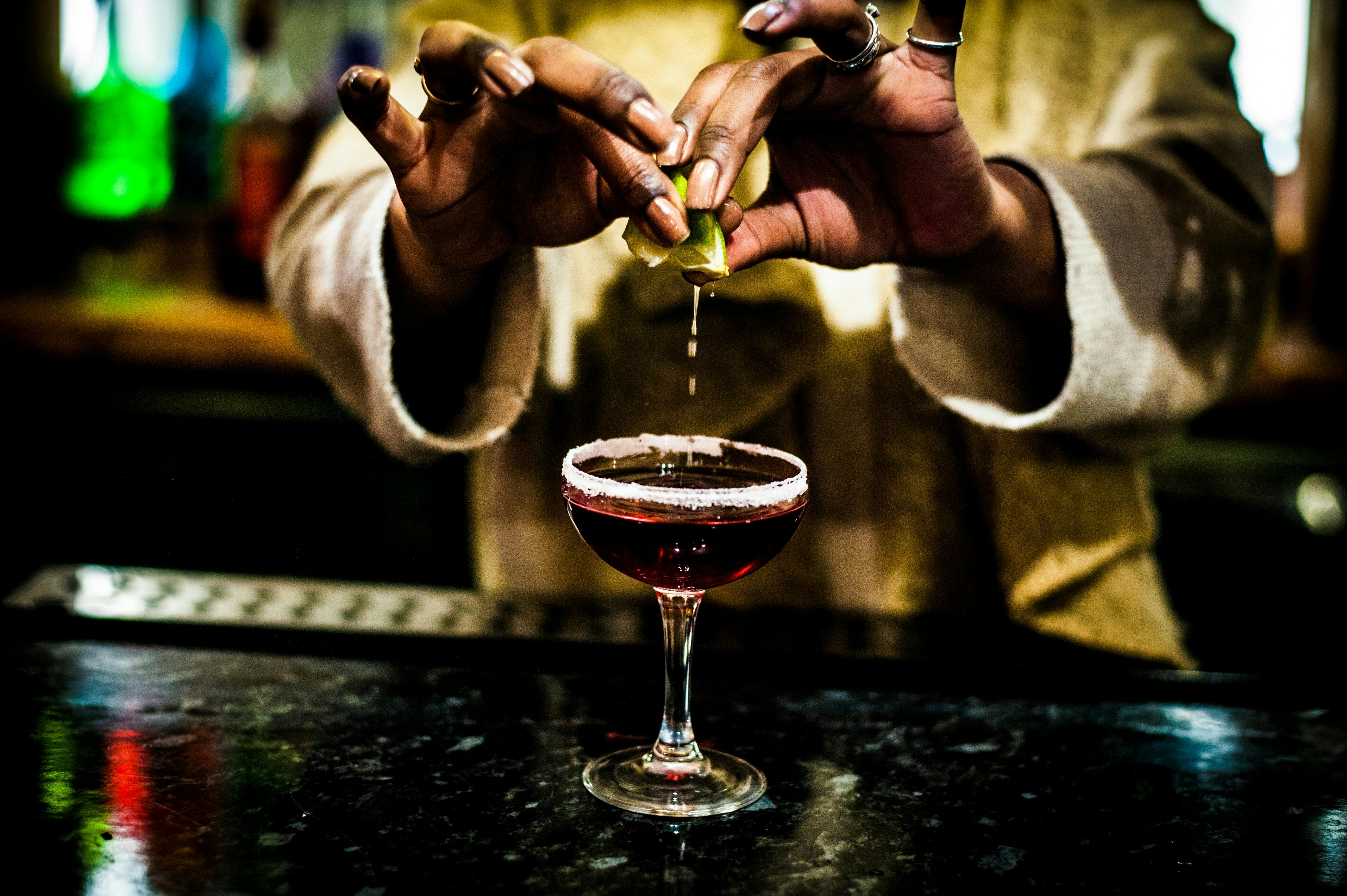 Close-up of a mixologist's hands squeezing a fresh lime wedge over a dark red cocktail in a sugar-rimmed coupe glass, with droplets of juice frozen in mid-air against a moody, blurred bar background.