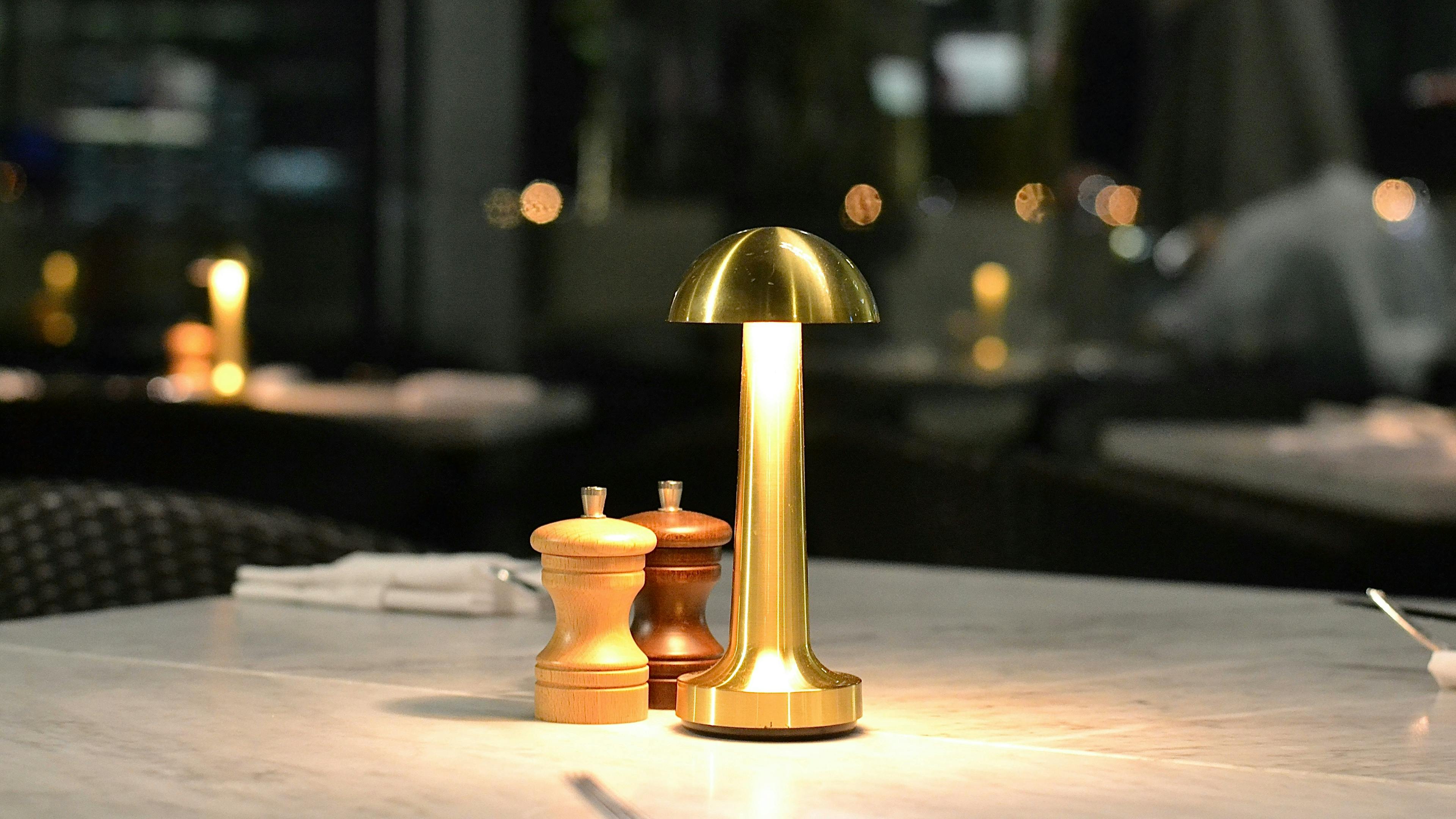 A minimalist gold cordless table lamp illuminating a white marble restaurant table at night, positioned next to light and dark wood salt and pepper shakers with a blurred dining room in the background.