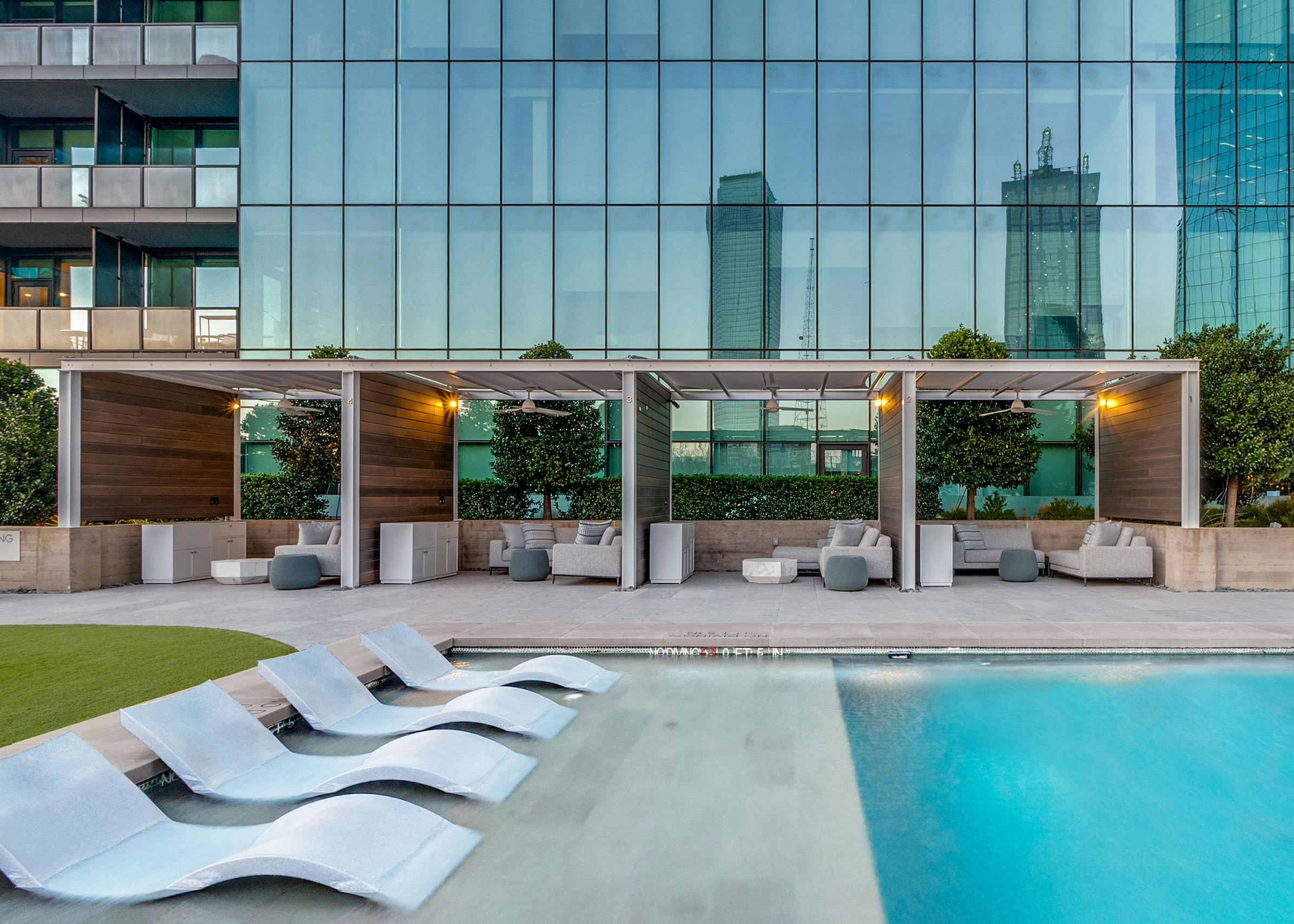 View of white sunset pool loungers and cabanas with furniture at AMLI Fountain Place with blue glass tower with windows in the background.