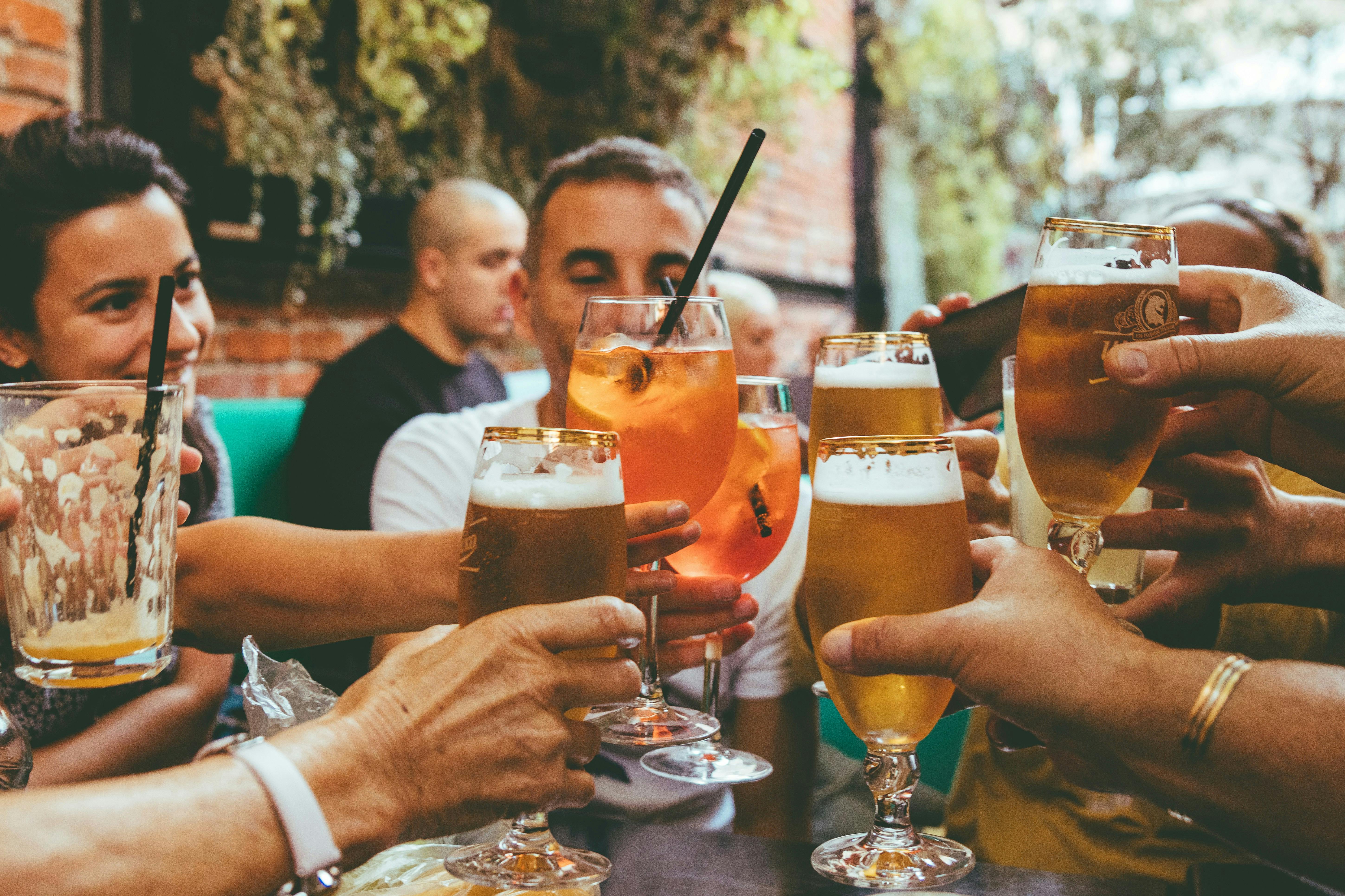 Close-up of a group of friends clinking glasses in a celebratory "cheers" at an outdoor bar. The variety of drinks includes tall glasses of golden beer and vibrant orange Aperol spritzes with black straws.