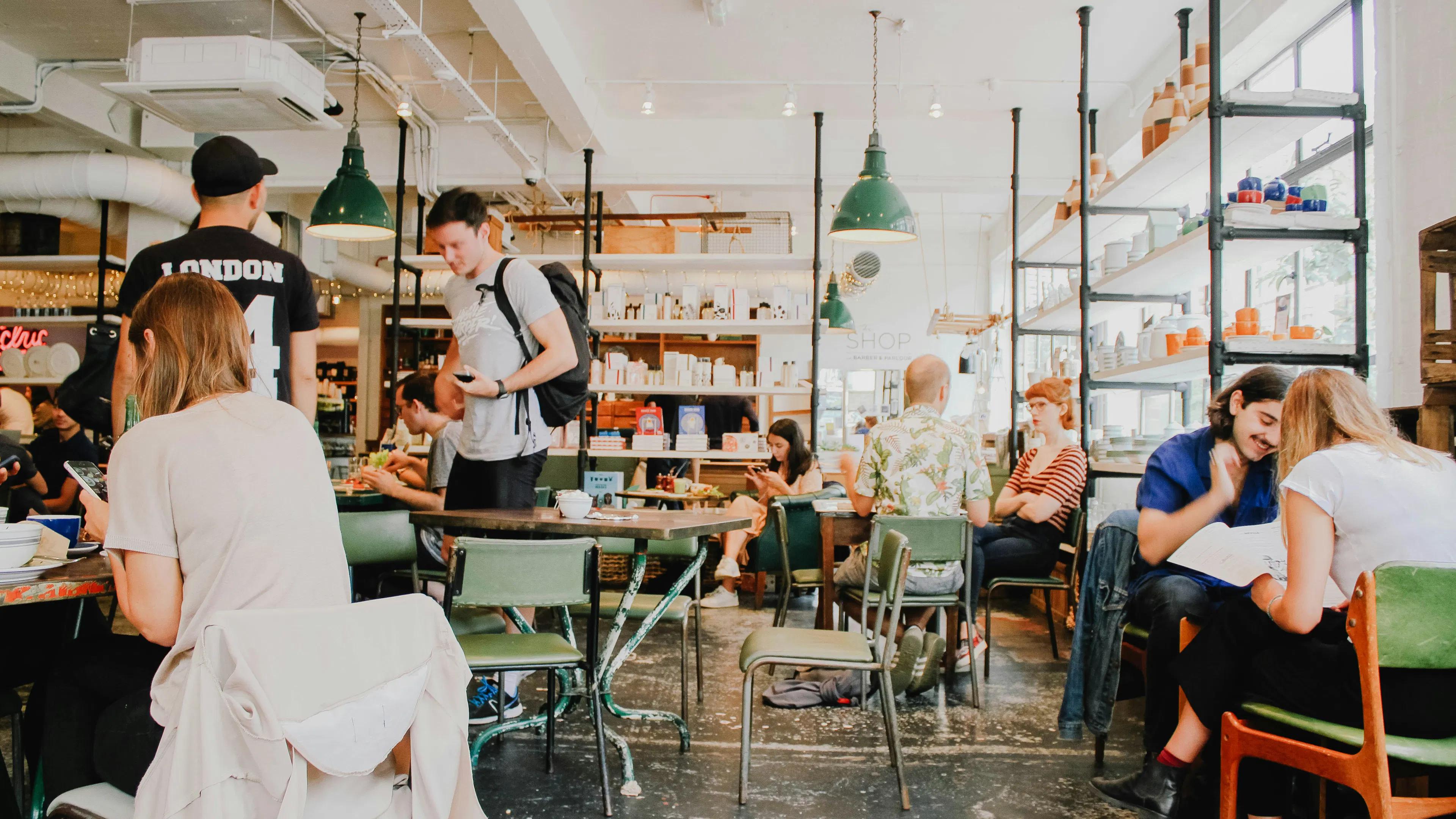 A bright, airy community cafe with white walls, high ceilings, and green industrial pendant lights. Customers are seated at various tables, while shelves along the wall display books and pottery, creating a vibrant social space.